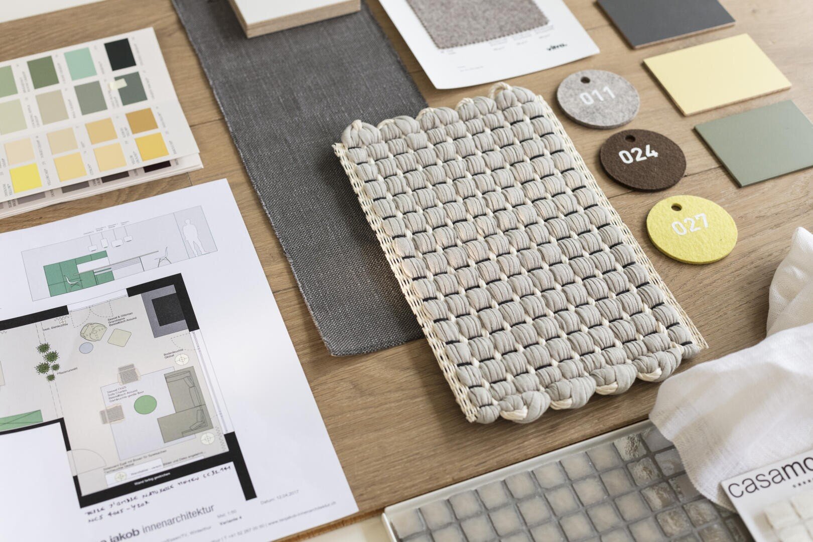 Des matériaux pour l'aménagement intérieur sont exposés sur une table : Des échantillons de tissus, des échantillons de couleurs, une petite natte tissée, des plans architecturaux et une palette de tons neutres et jaunes, clairement disposés pour une présentation de projet.