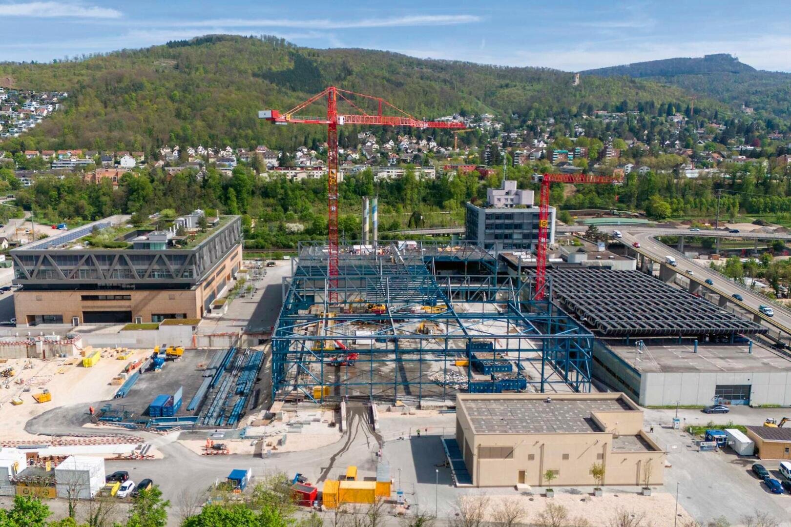 Luftaufnahme einer Großbaustelle mit Kränen, Stahlgerüsten und mehreren Gebäuden, umgeben von Bäumen, Hügeln und einem Wohngebiet im Hintergrund bei klarem Himmel.