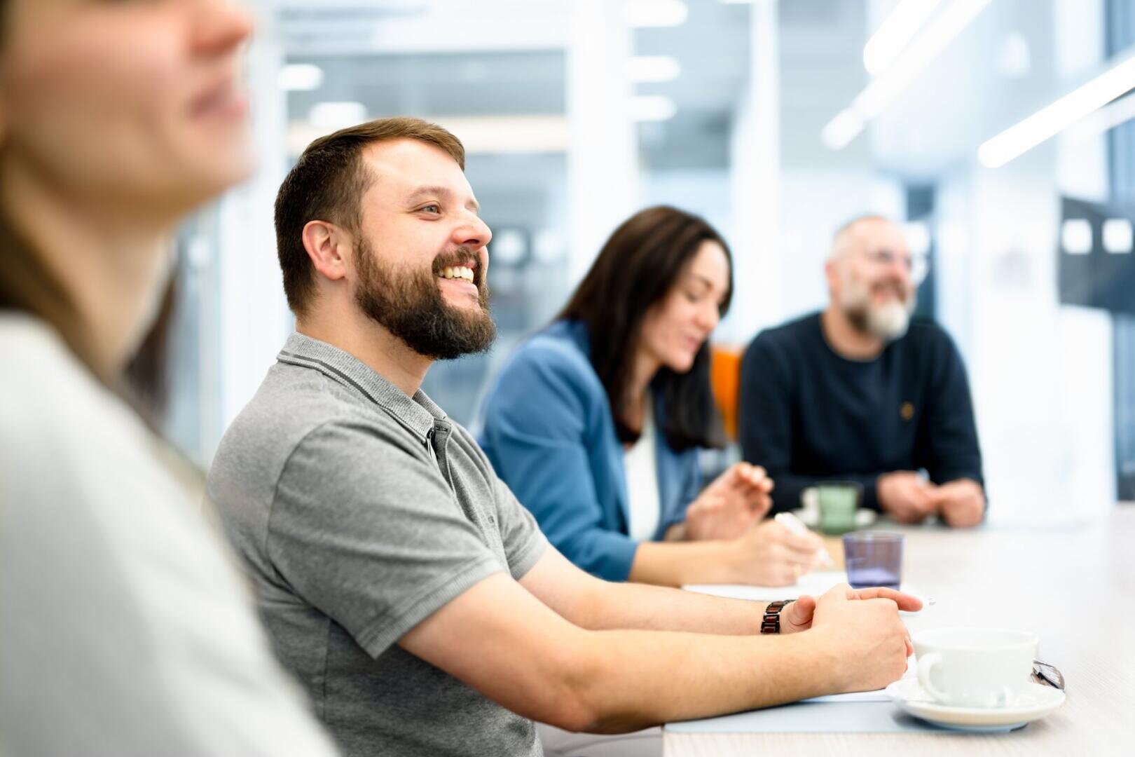 Eine Gruppe von vier Erwachsenen sitzt an einem Tisch in einem hellen, modernen Büro, lächelt und unterhält sich während einer Besprechung. Ein Mann im Vordergrund lacht, während die anderen aufmerksam und entspannt wirken.