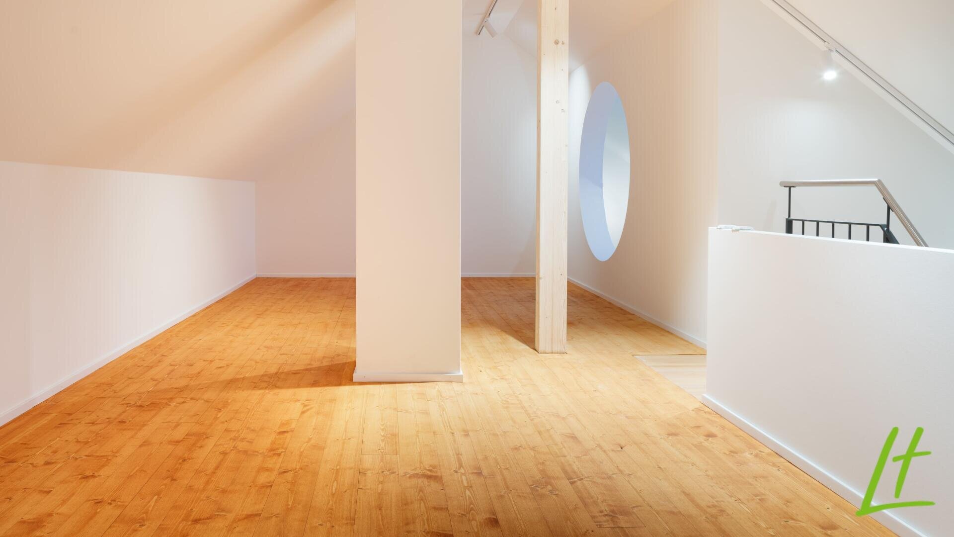 Ein heller, moderner Loft-Raum mit hellem Holzfußboden, weißen Wänden, einer zentralen Stützsäule, einem hohen Holzbalken, einem runden Fenster und einem minimalistischen Geländer auf der rechten Seite. Das grüne "Lt"-Logo erscheint in der unteren rechten Ecke.