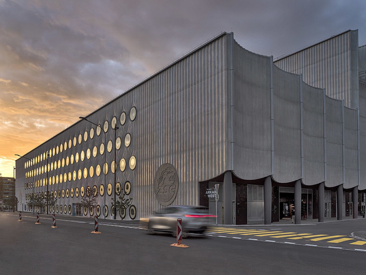 Ein modernes, graues Gebäude mit einer Fassade aus runden Fenstern in einem Gittermuster, wellenförmigen Architekturelementen und Autos, die bei Sonnenuntergang auf einer Straße davor vorbeifahren.