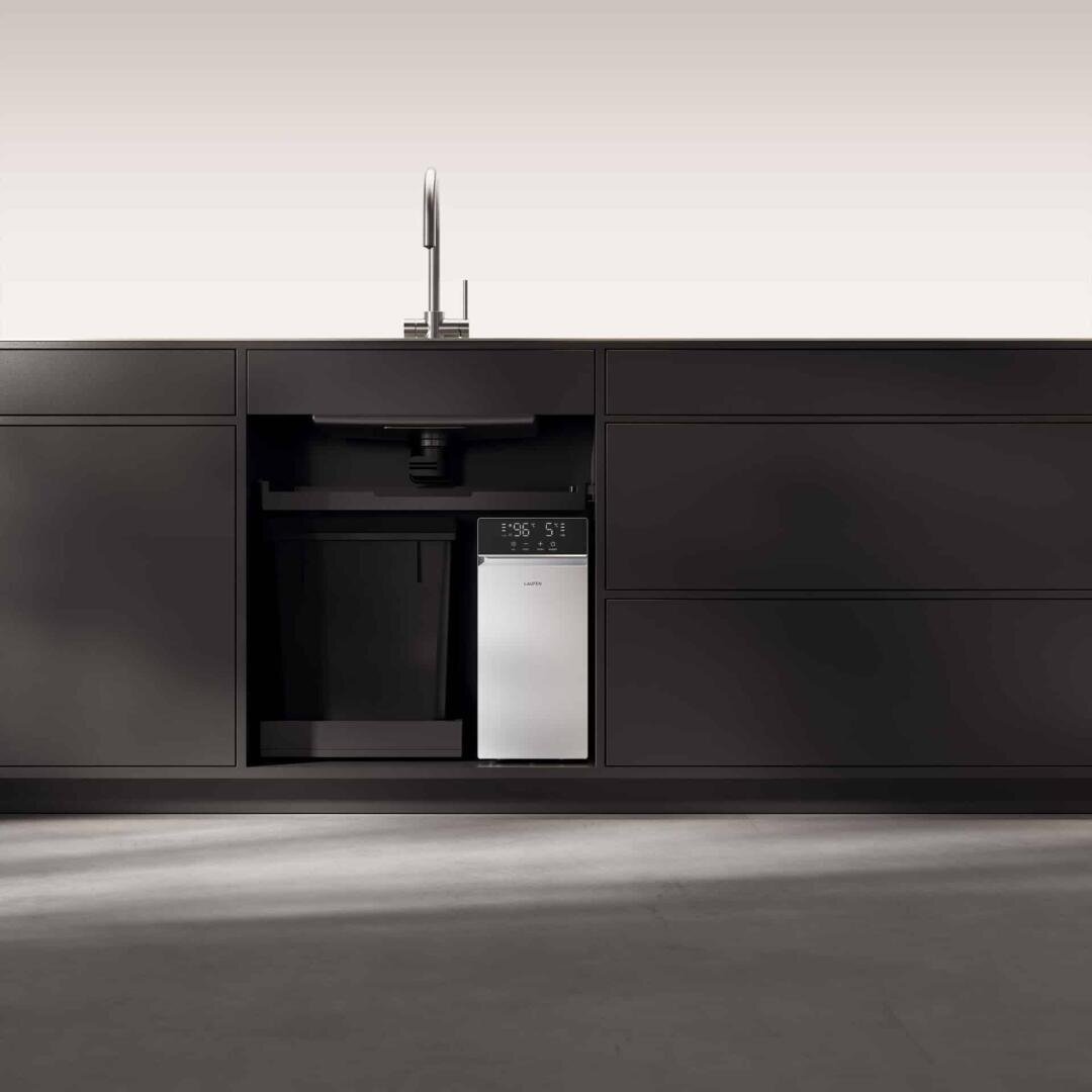 Minimalistische schwarze Küchenschränke mit einem modernen Spülbecken darüber. Unter der Spüle ist ein kompaktes, silbernes Gerät mit Digitalanzeige installiert, das von leeren Schränken umgeben ist. Der Boden ist schlicht und grau.