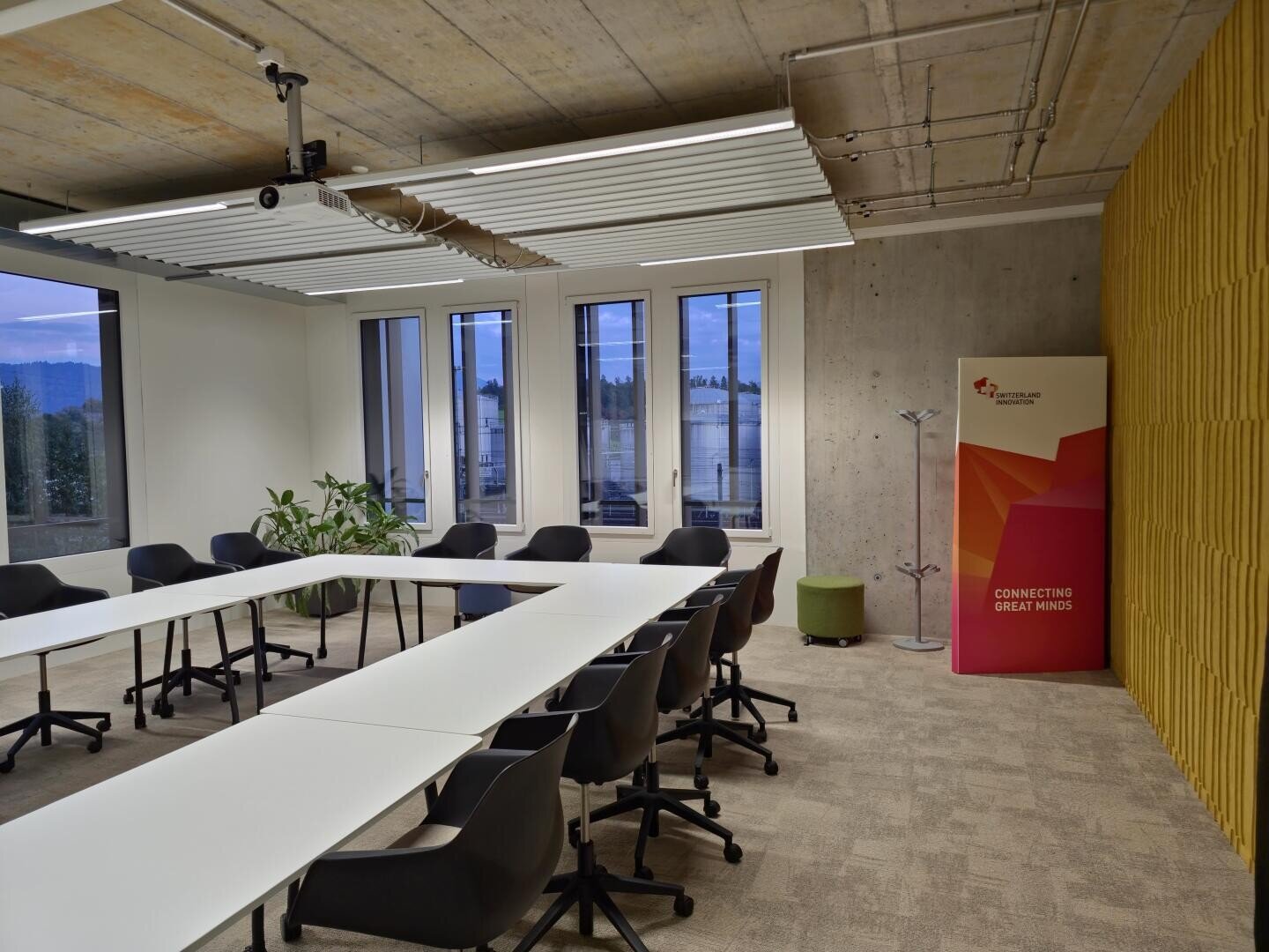 Une salle de conférence moderne avec une disposition en U de tables blanches et de chaises noires, de grandes fenêtres, des plantes, un projecteur et une bannière indiquant "Connecter les grands esprits" près d'un mur d'accent jaune.