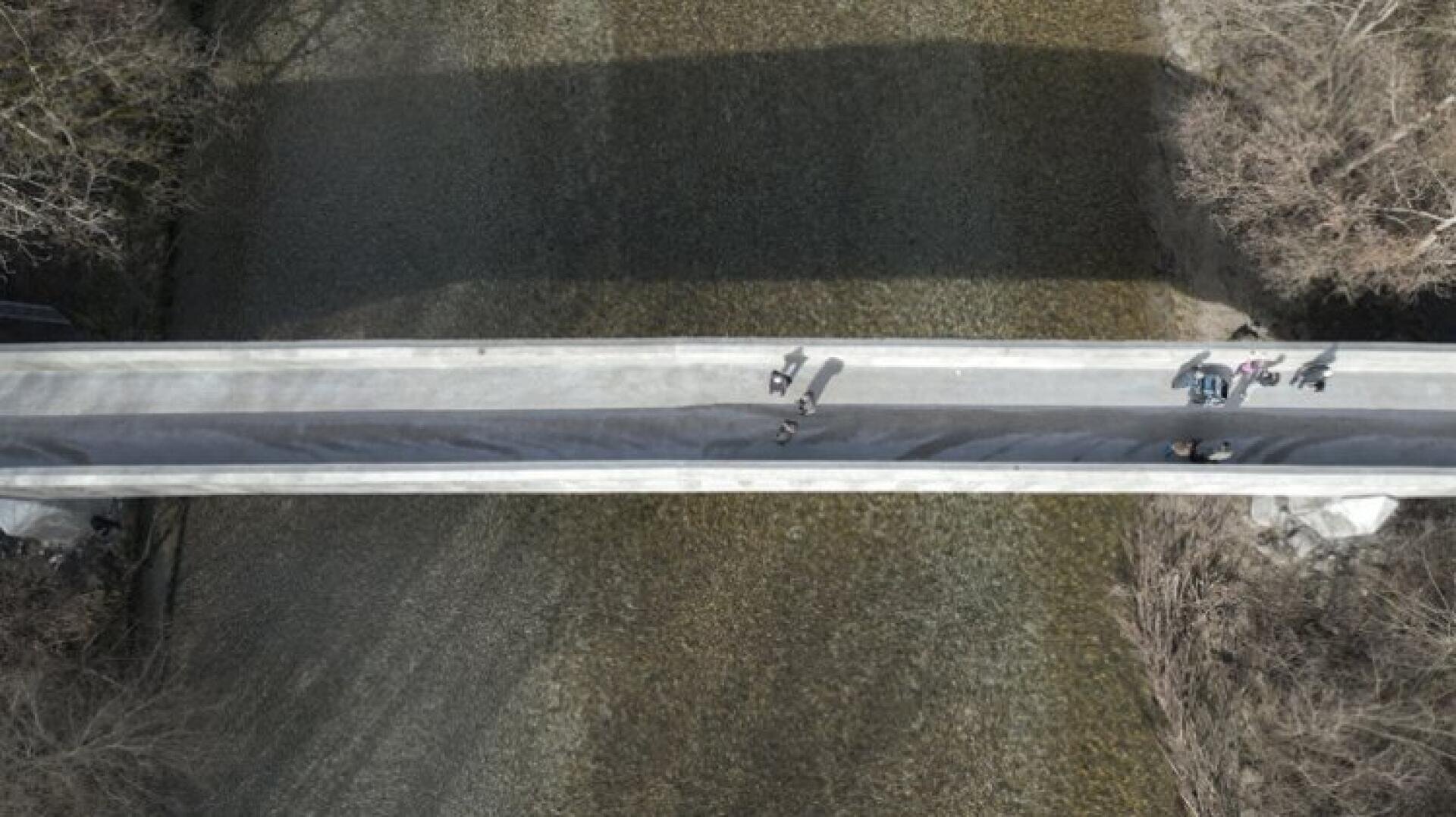 Luftaufnahme einer schmalen Brücke über einen seichten Fluss mit ein paar Fußgängern und Radfahrern und einigen Bäumen an beiden Flussufern, die Schatten auf das Wasser werfen.