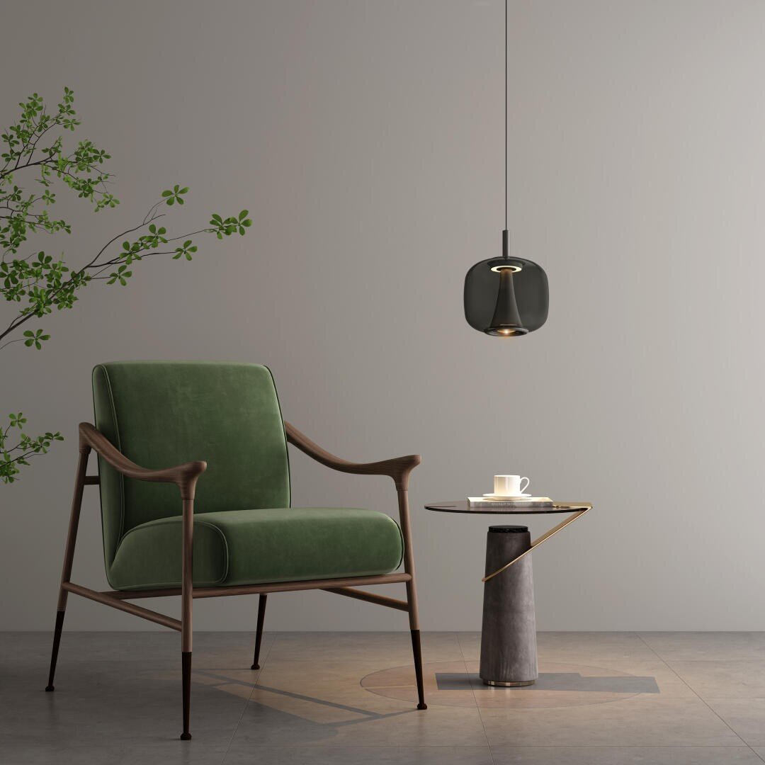 Ein modernes Interieur mit einem grünen Sessel, einem kleinen runden Beistelltisch mit Tasse und Untertasse, einer schwarzen Pendelleuchte darüber und einer blattreichen Pflanze vor einer schlichten grauen Wand.