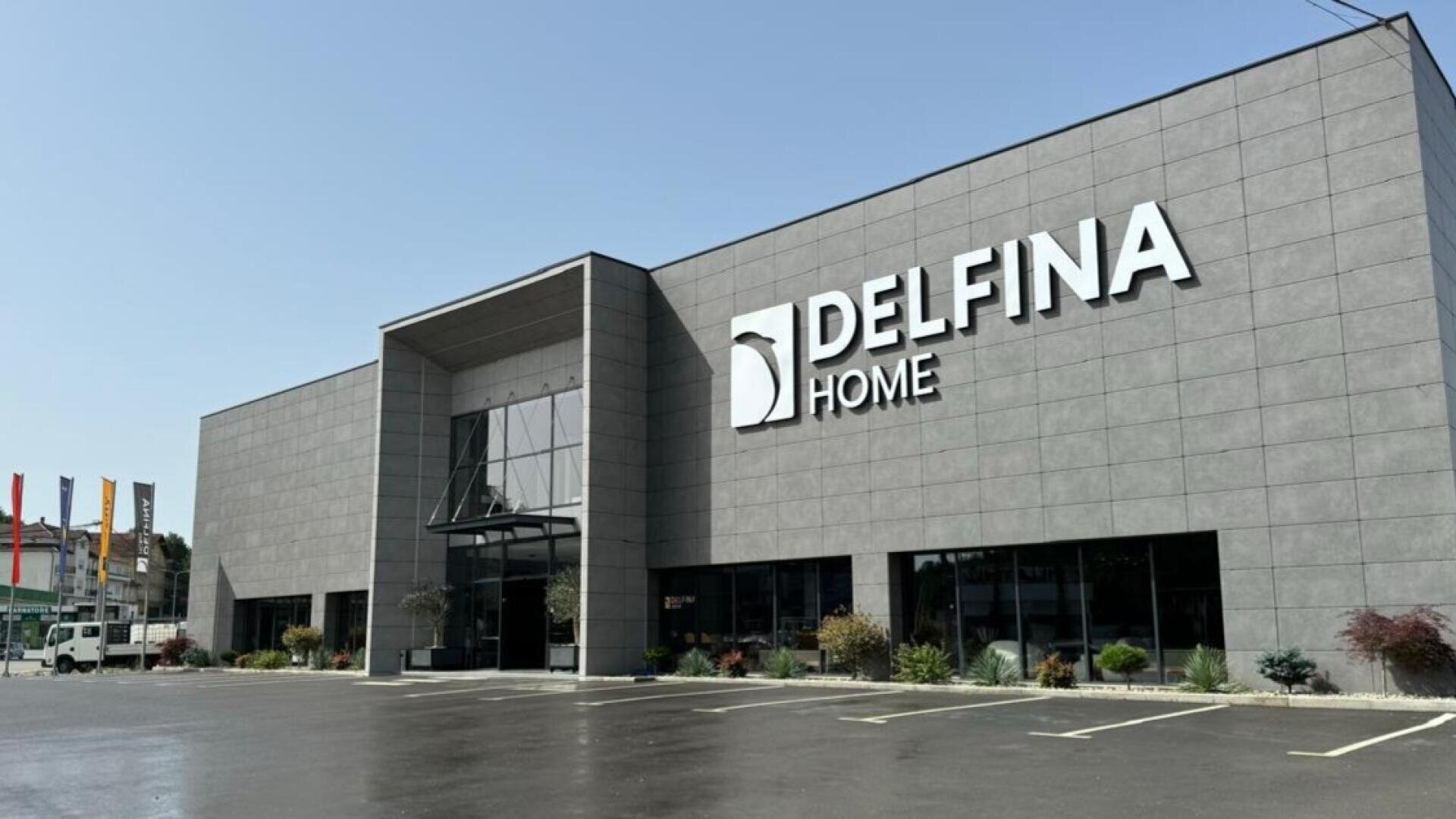 Ein modernes graues Gebäude mit großen Fenstern und einem Schild mit der Aufschrift DELFINA HOME an der Fassade. Der Parkplatz davor ist fast leer, und um den Eingang sind einige kleine Bäume und Sträucher gepflanzt.