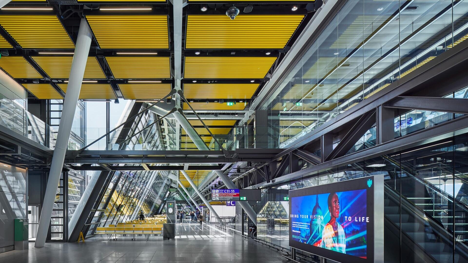 Ein modernes, geräumiges Flughafenterminal mit gelben Deckenplatten, Glaswänden, Metallträgern, Rolltreppen und einer digitalen Werbetafel. Sonnenlicht strömt herein, und im Hintergrund sind Schilder und leere Sitzplätze zu sehen.
