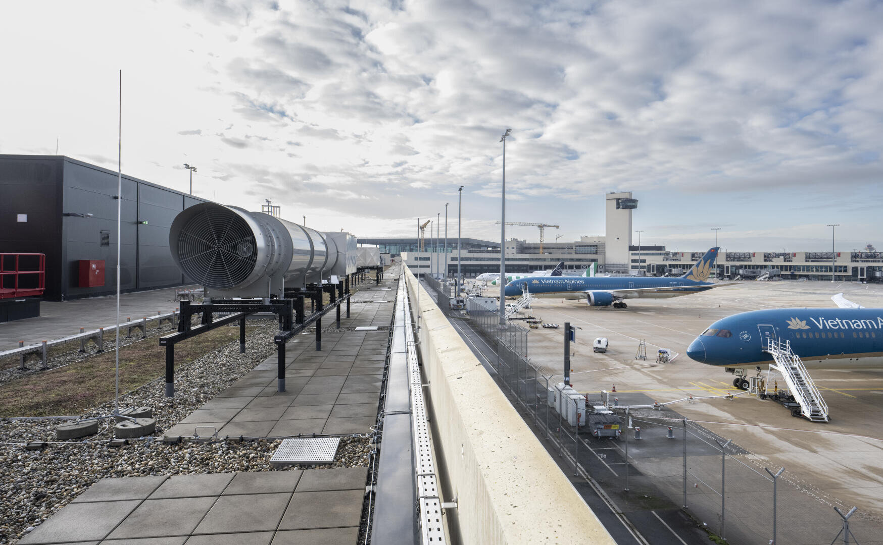 Blick auf das Vorfeld eines Flughafens mit Flugzeugen der Vietnam Airlines, die an den Gates geparkt sind, Bodenservicefahrzeugen in der Nähe und einem Kontrollturm im Hintergrund bei teilweise bewölktem Himmel. Im Vordergrund sind Lüftungsanlagen auf dem Dach zu sehen.