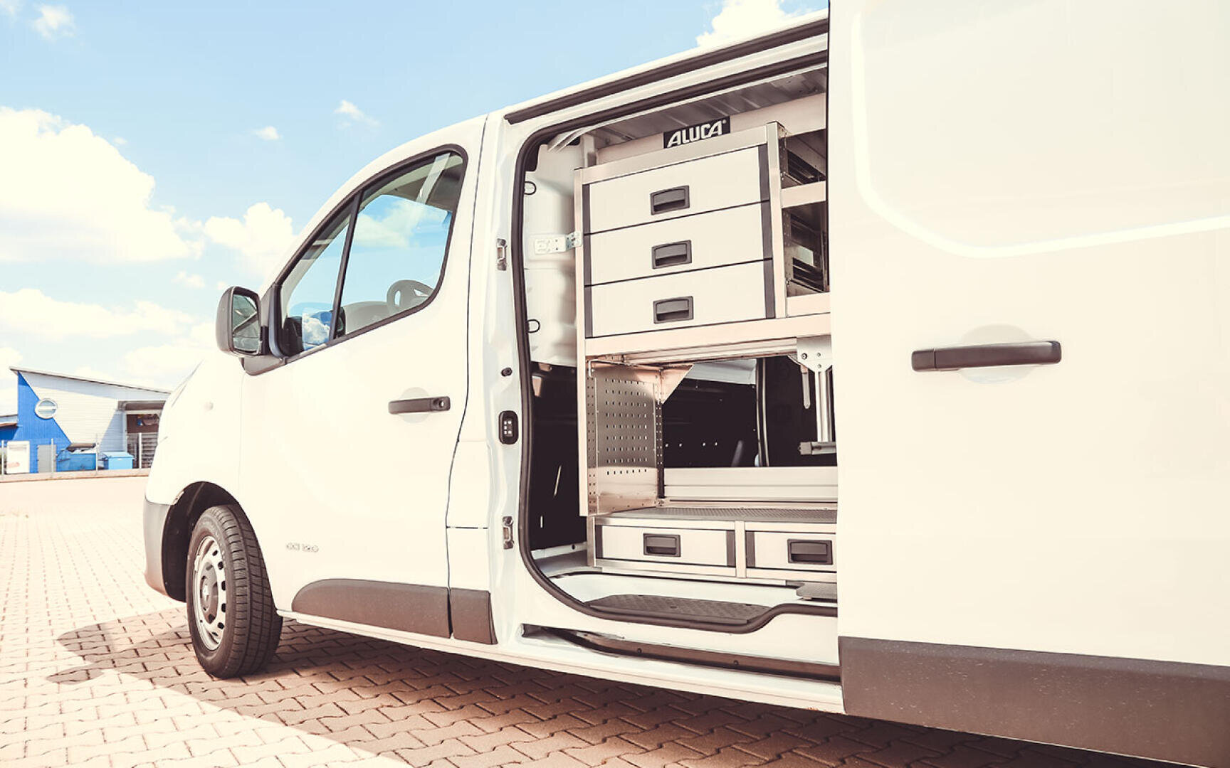 Ein weißer Lieferwagen, dessen Seitentür geöffnet ist, gibt den Blick auf geordnete Metallschubladen und Regale frei, die für Werkzeuge oder Ausrüstung gedacht sind. Der Lieferwagen ist auf einer gepflasterten Fläche unter einem teilweise bewölkten Himmel geparkt.