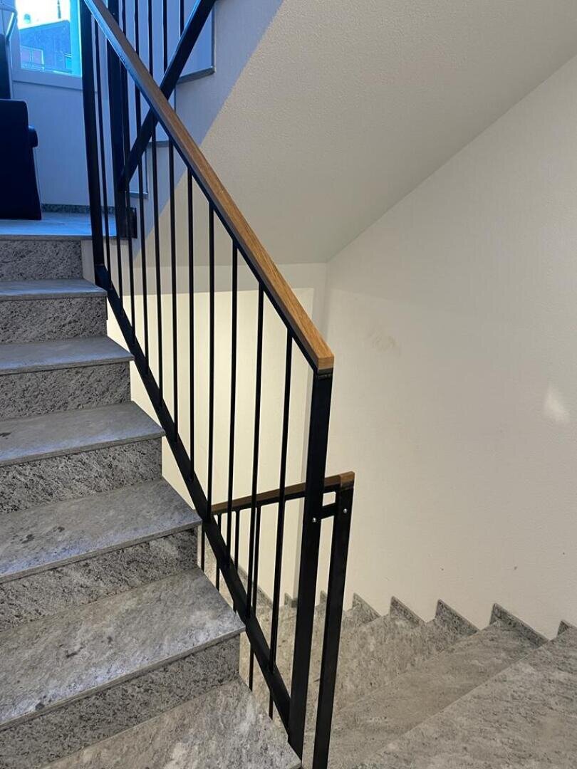 Une cage d'escalier avec des marches en pierre grise, une rampe métallique noire avec une main courante en bois et des murs blancs. L'escalier bifurque à un palier et mène aussi bien vers le haut que vers le bas. La lumière naturelle entre par une fenêtre située à proximité.
