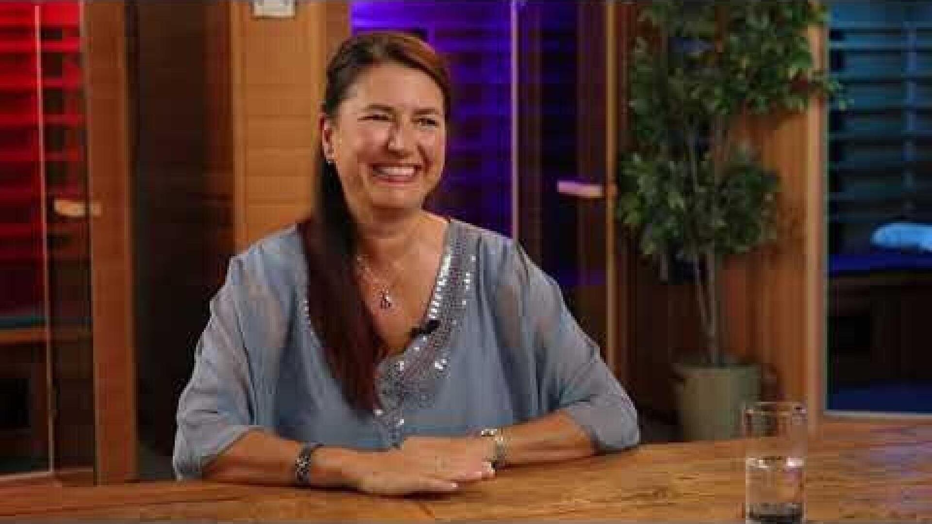 Eine Frau mit langen braunen Haaren und einer hellblauen Bluse sitzt lächelnd an einem Holztisch. Auf dem Tisch steht ein Glas Wasser, im Hintergrund sind bunte Lichter und eine Topfpflanze zu sehen.