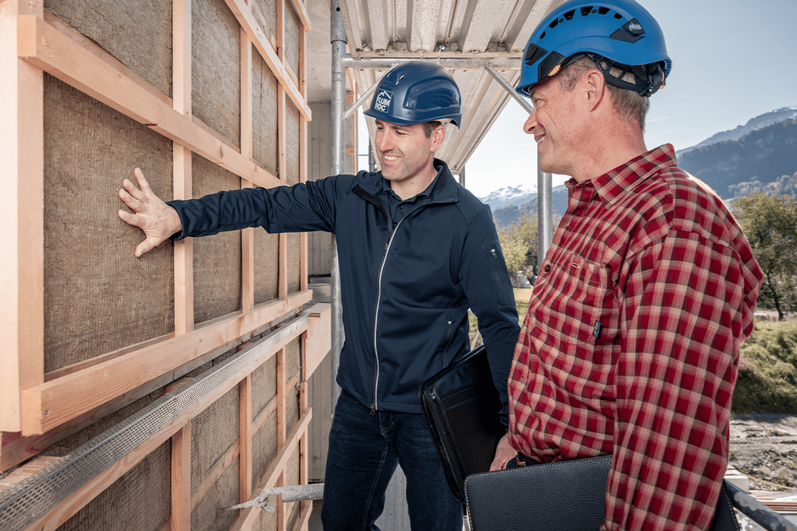 Deux hommes portant un casque bleu inspectent l'isolation extérieure d'un bâtiment en construction. Un homme gesticule en direction du mur, tandis que l'autre, vêtu d'une chemise à carreaux, tient un dossier noir. En arrière-plan, on aperçoit des montagnes.