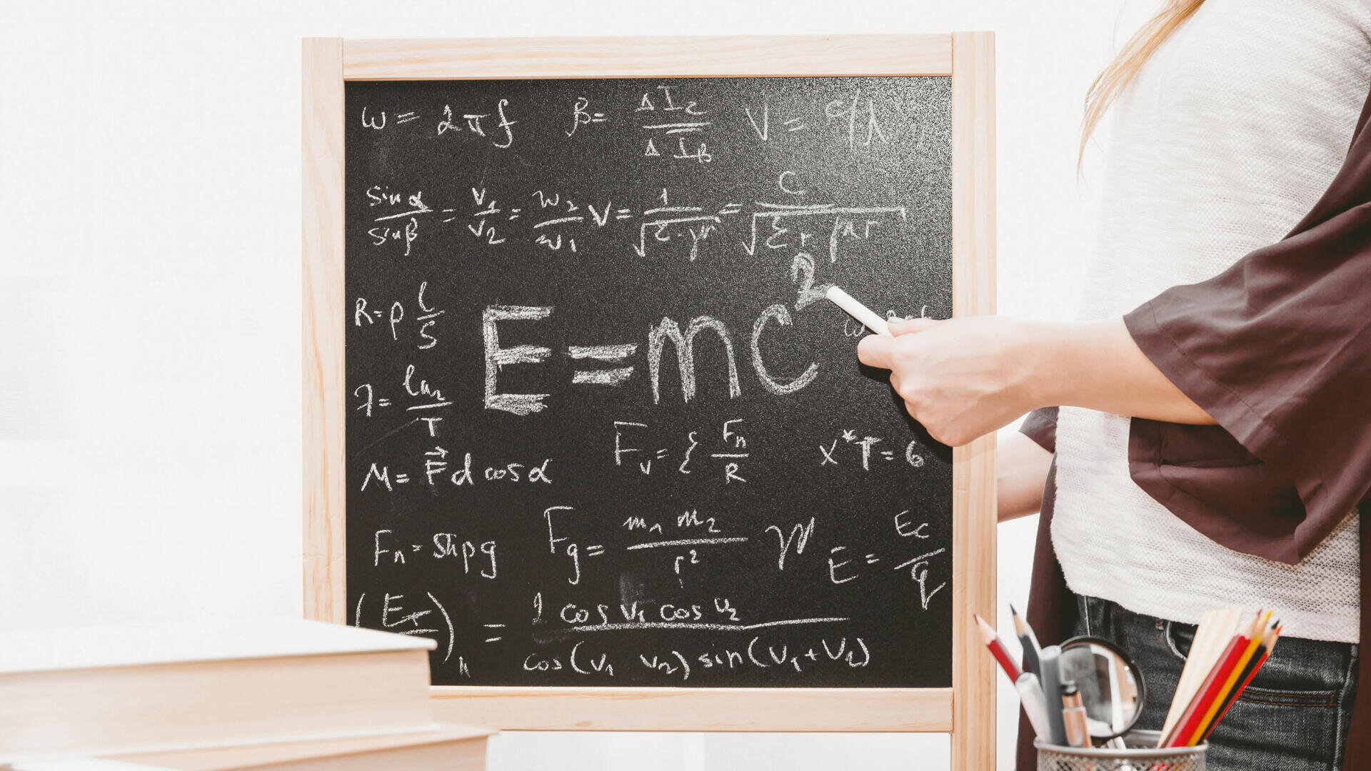 Eine Person steht vor einer Tafel mit physikalischen Formeln, darunter Einsteins Gleichung E=mc², und hält ein Stück Kreide. In der Nähe befinden sich Bücher und ein Becher mit Stiften.