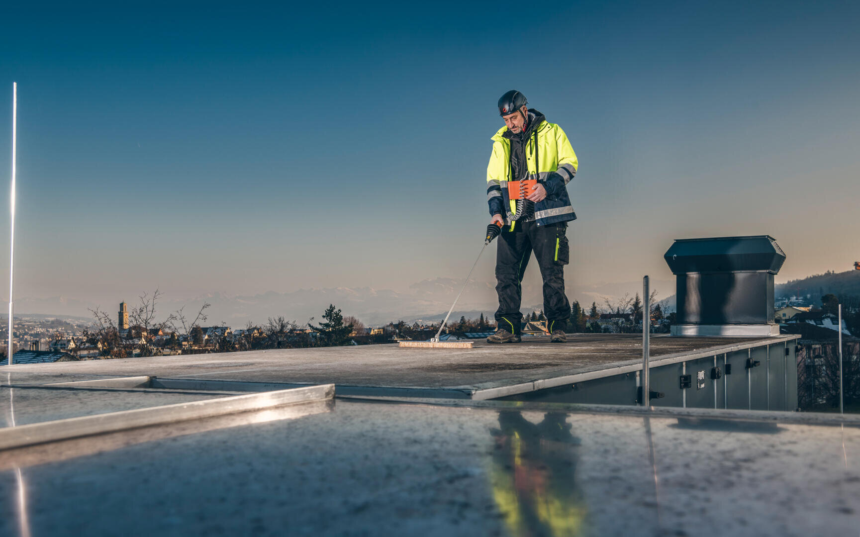 Ein Arbeiter in Warnweste und Schutzhelm steht auf einem Dach und arbeitet mit einem Werkzeug. Im Hintergrund sind bei klarem Himmel die Gebäude der Stadt und die Berge zu sehen.