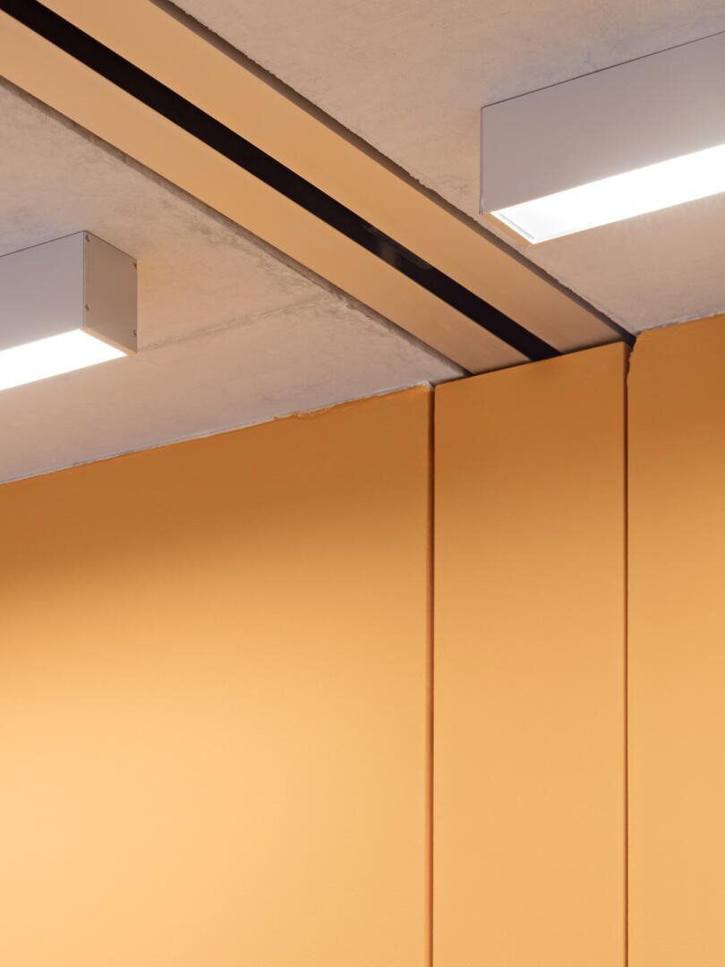 Minimalistisches Interieur mit einer gelb-orangenen Wand, die auf eine Betondecke trifft, mit zwei rechteckigen Deckenleuchten und dunklen Linien, die die Fugen zwischen den Oberflächen markieren. Das Design ist klar und geometrisch.