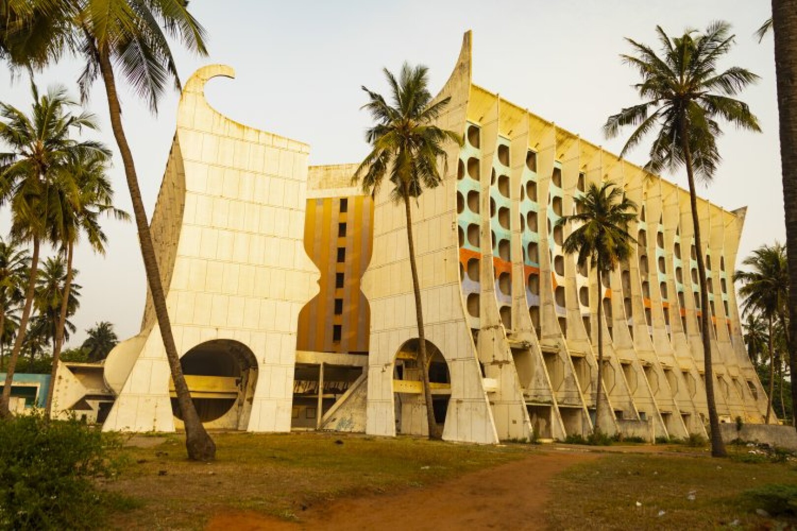 Ein einzigartiges, modernistisches Gebäude mit geometrischen Formen und farbenfrohen Akzenten, umgeben von hohen Palmen und einem Feldweg, unter klarem Himmel. Die Architektur zeichnet sich durch Rundbögen und eine markante, skulpturale Fassade aus.