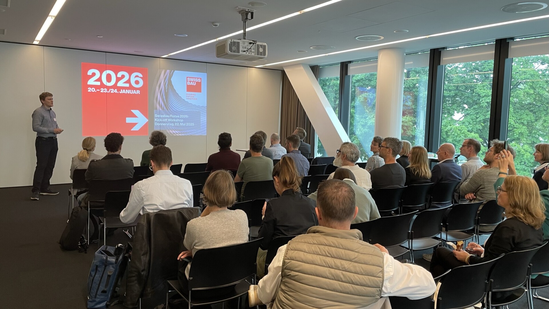 Ein Mann steht vor einem modernen Konferenzraum und hält einen Vortrag vor einem sitzenden Publikum. Auf einem Bildschirm werden „20.–23. Januar 2026“ und weitere Veranstaltungsdetails angezeigt. Große Fenster geben den Blick auf Bäume draußen frei.