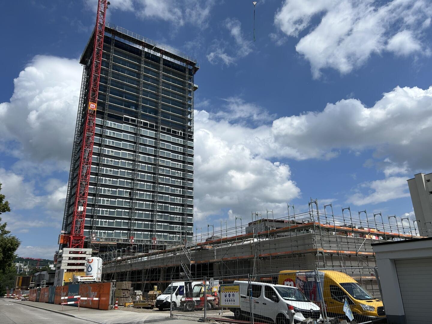 Ein im Bau befindliches Hochhaus mit Gerüst und rotem Kran, daneben ein niedrigeres Gebäude. Mehrere Lieferwagen und Baumaterialien sind auf der Baustelle unter einem teilweise bewölkten Himmel zu sehen.