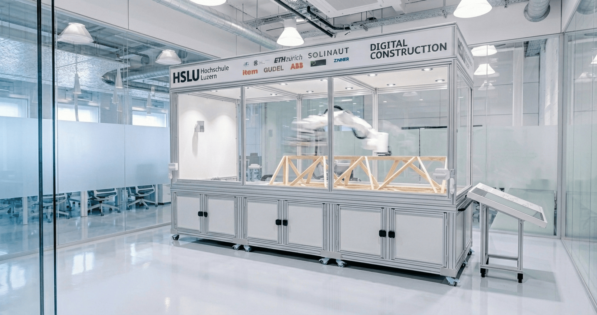 Ein Roboterarm konstruiert Holzgerüste in einem gläsernen Labor der Hochschule Luzern, mit Schildern für Partnerorganisationen darüber und einem Touchscreen-Bedienfeld auf der rechten Seite. Der moderne Raum ist hell und sauber.