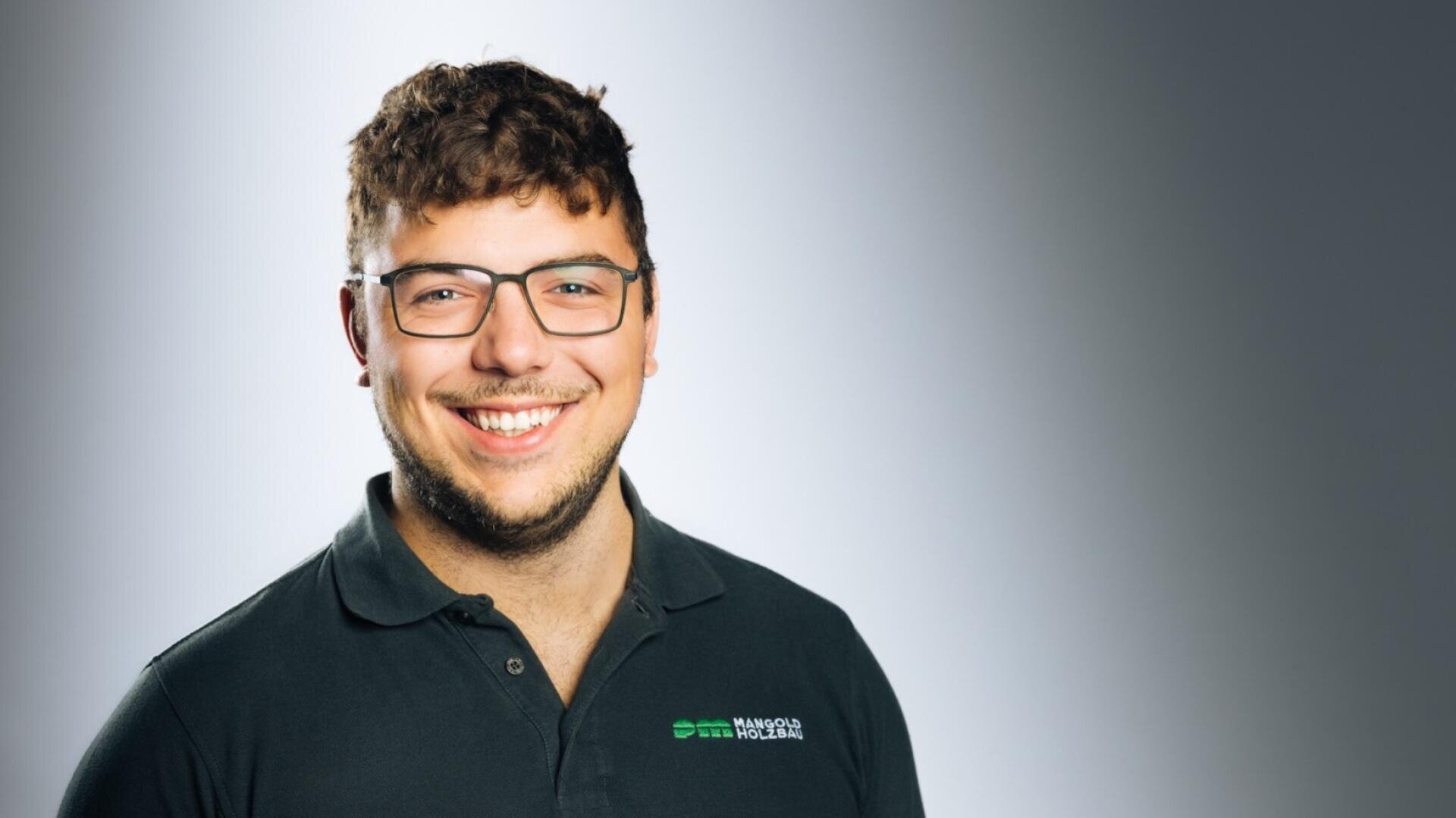 Ein junger Mann mit kurzen, gelockten braunen Haaren und einer Brille lächelt, während er ein dunkles Polohemd mit einem kleinen grün-weißen Logo auf der linken Brust vor einem Hintergrund mit grauem Farbverlauf trägt.