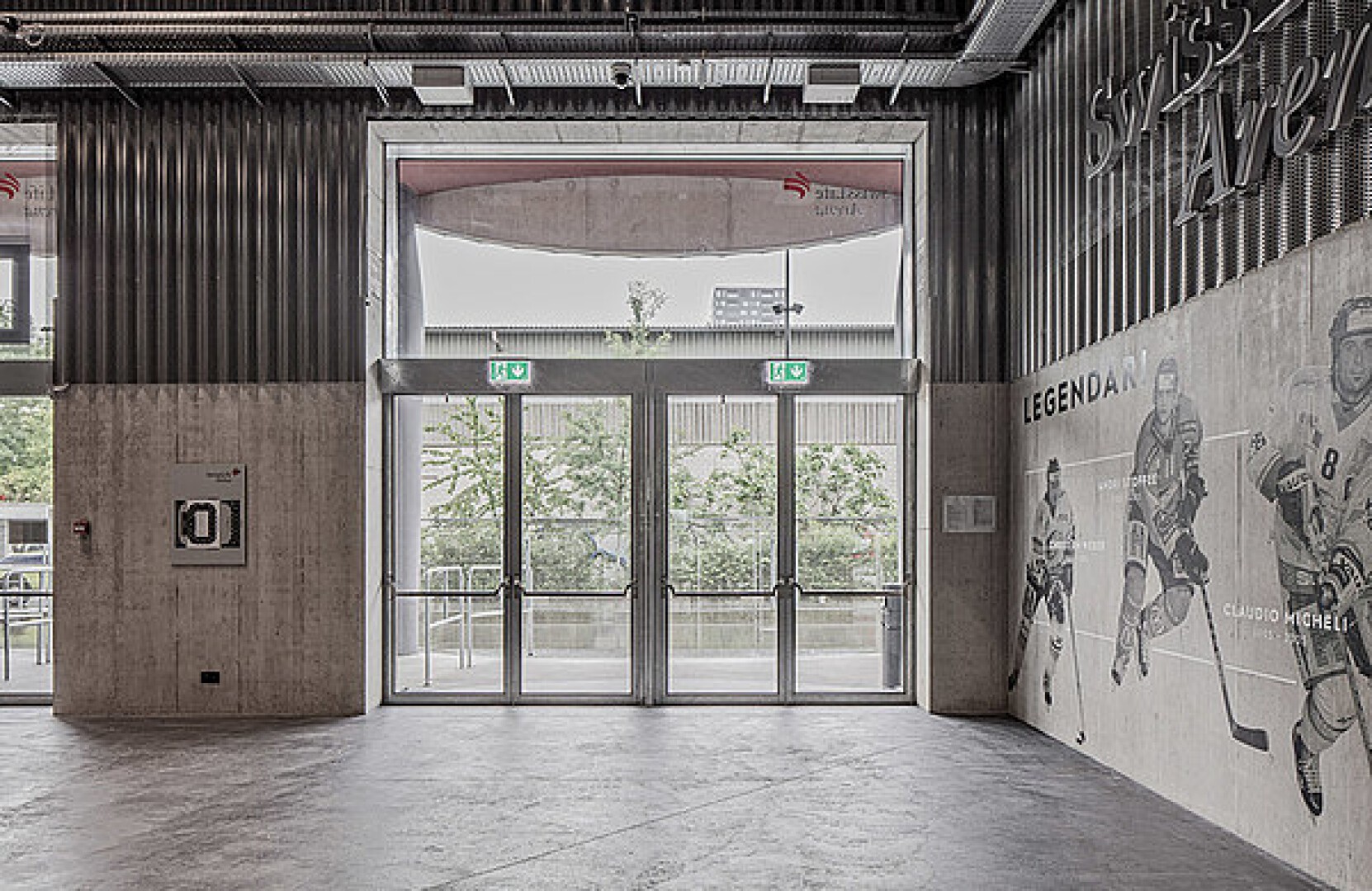 Ein moderner Gebäudeeingang mit Doppelglastüren, darüber angebrachten „EXIT“-Schildern und einem großen Sportwandbild mit Hockeyspielern an der rechten Wand. Durch die Türen sind Bäume und ein Außenbereich sichtbar.