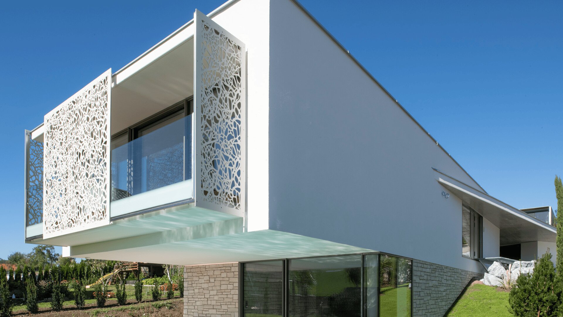Modernes weißes Haus mit großen Glasfenstern und dekorativen perforierten Metallpaneelen im Obergeschoss, mit einer freitragenden Konstruktion und einem Steinsockel, vor einem klaren blauen Himmel.