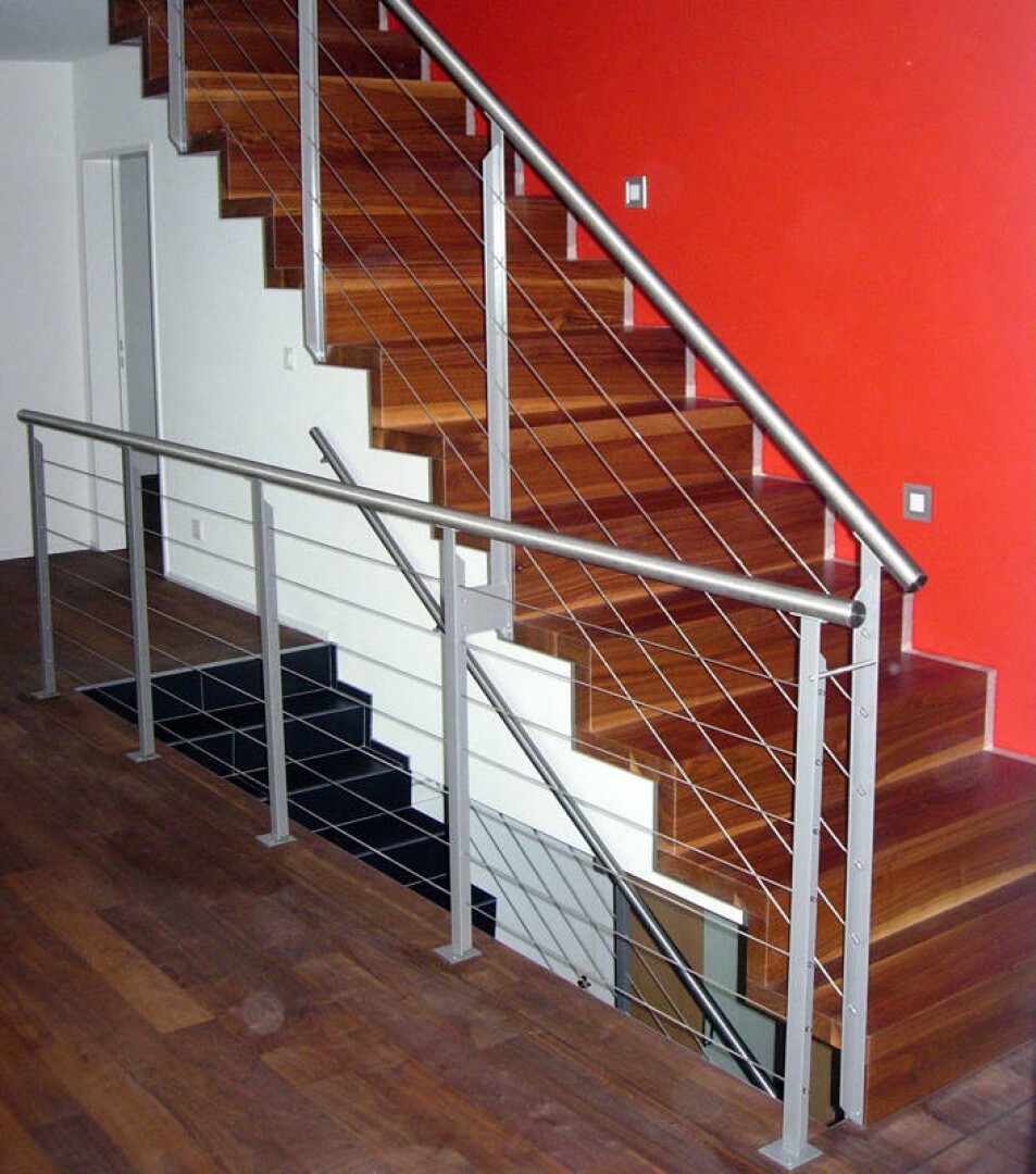 Moderne Innentreppe mit Holzstufen, einem Edelstahlgeländer mit horizontalen Kabeln und einer leuchtend roten Akzentwand. Die Treppe hat ein eckiges, geometrisches Design und befindet sich über einer niedrigeren Treppe.
