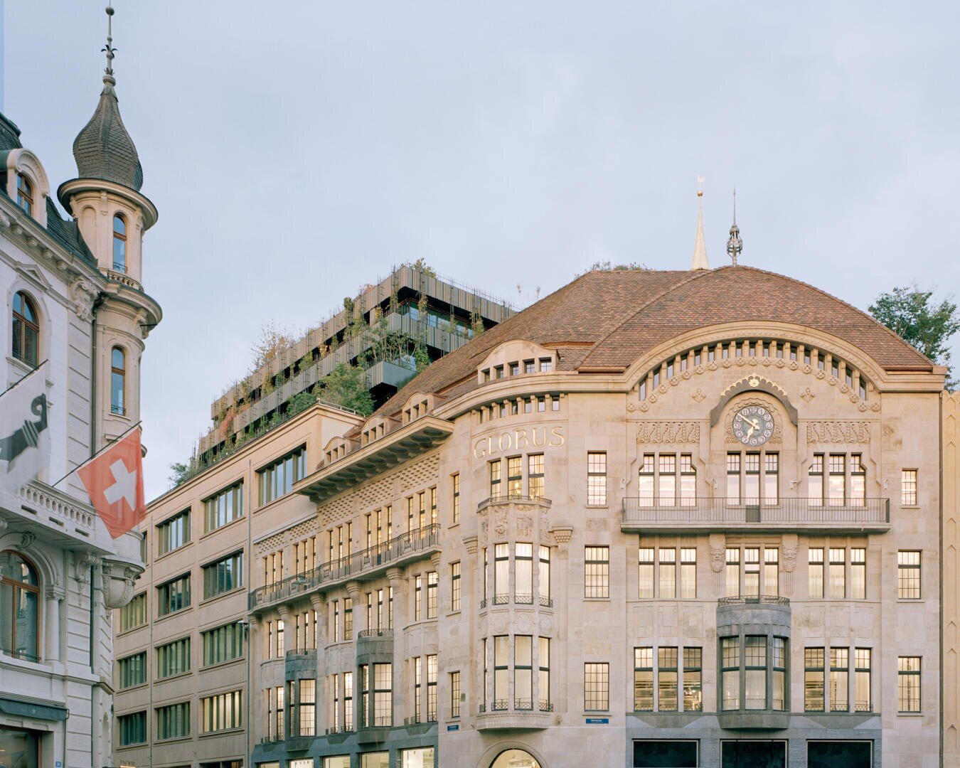 In einer Stadt steht ein historisches Steingebäude mit verschnörkelten architektonischen Details und einer Uhr über dem Eingang. Daneben hängt eine Schweizer Flagge an einem anderen Gebäude mit einem Türmchen. Im Hintergrund sind moderne Strukturen mit Grünflächen zu sehen.