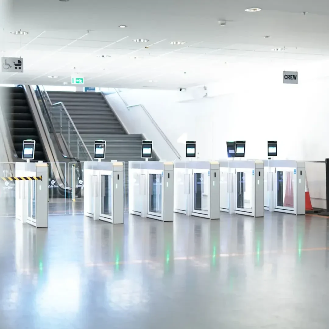 Eine Reihe von automatischen Sicherheitsschleusen mit digitalen Bildschirmen in einem modernen, leeren Flughafenterminal. Im Hintergrund sind eine Rolltreppe und ein Schild mit der Aufschrift CREW zu sehen.