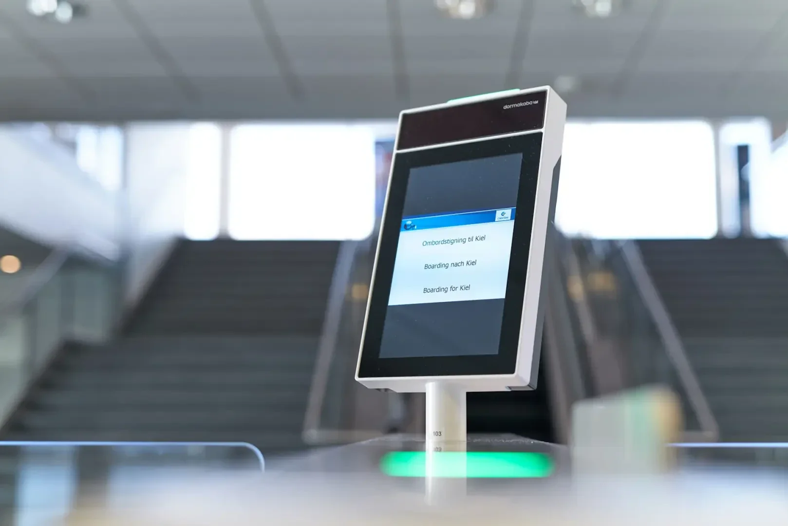 Eine Nahaufnahme eines elektronischen Flugsteig-Kiosks mit einem Bildschirm, der Informationen zum Einsteigen anzeigt, in der Nähe einer Rolltreppe und einer Treppe in einer hellen, modernen Flughafenumgebung.