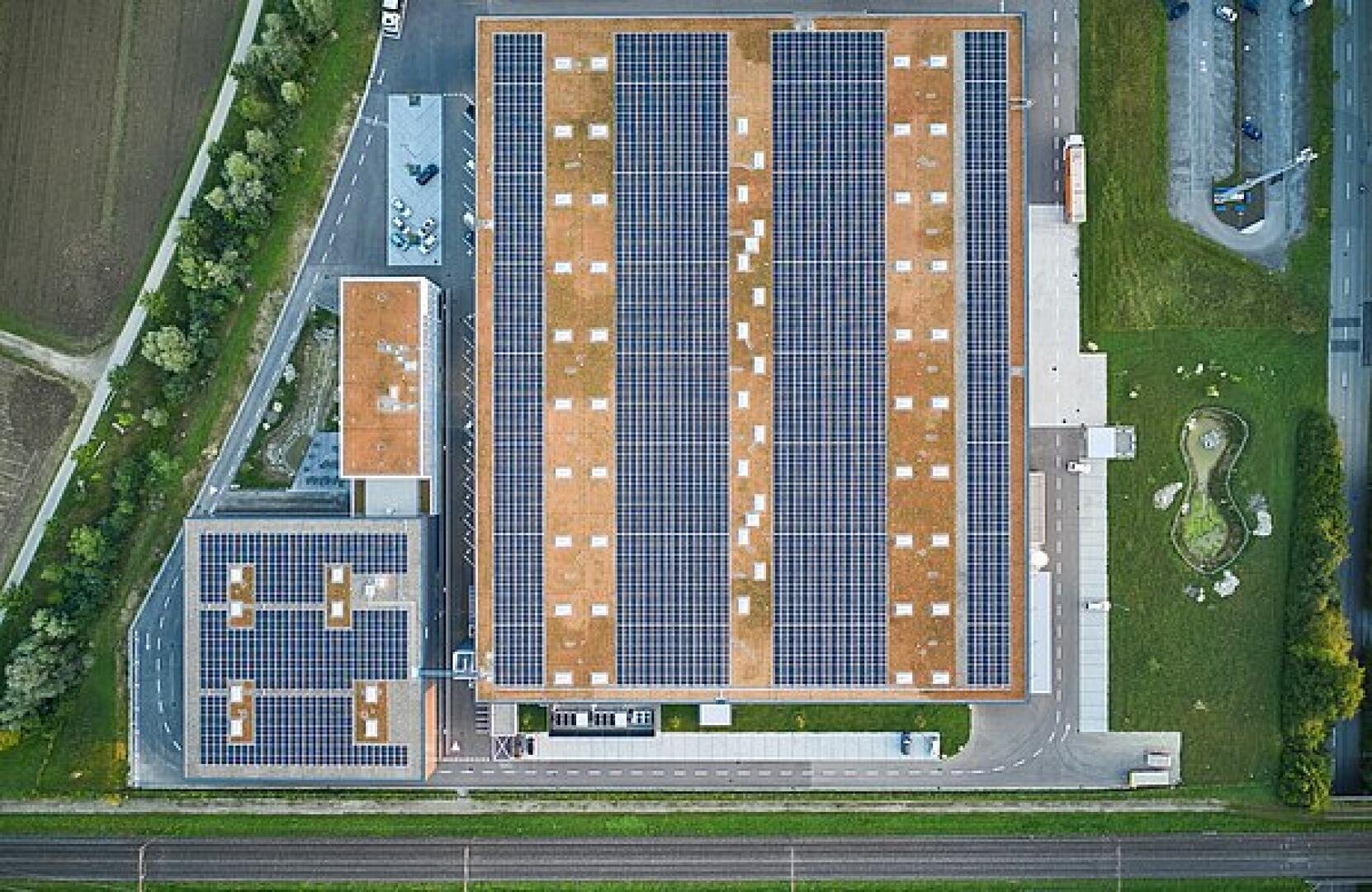 Luftaufnahme von zwei großen Gebäuden mit Sonnenkollektoren auf den Dächern, umgeben von Gras, Straßen und einem Teich, die die nachhaltige Energienutzung in einem Industriegebiet veranschaulichen.