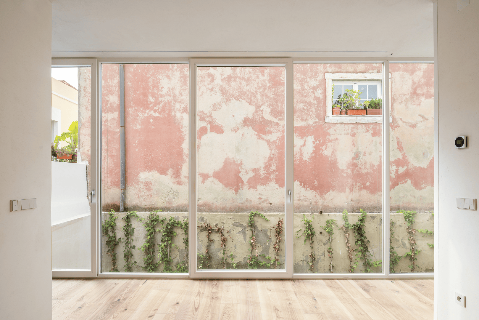 Heller Innenraum mit raumhohen Glastüren und Blick auf einen kleinen Innenhof mit Grünpflanzen und einer verwitterten, rosafarbenen Wand mit einem Fenster und einem Blumenkasten draußen. Heller Holzboden und minimale Ausstattung im Inneren.