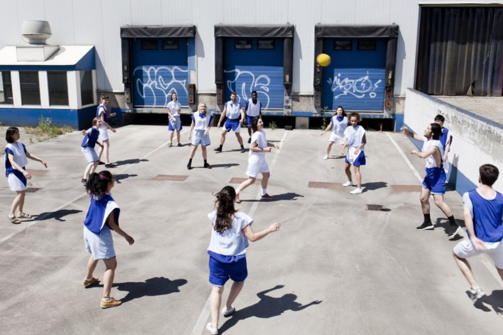 Eine Gruppe von Teenagern in blau-weißen Sportuniformen spielt auf einem gepflasterten Außengelände, umgeben von Industriegebäuden mit Graffiti auf den Garagentoren, ein Spiel mit einem gelben Ball.