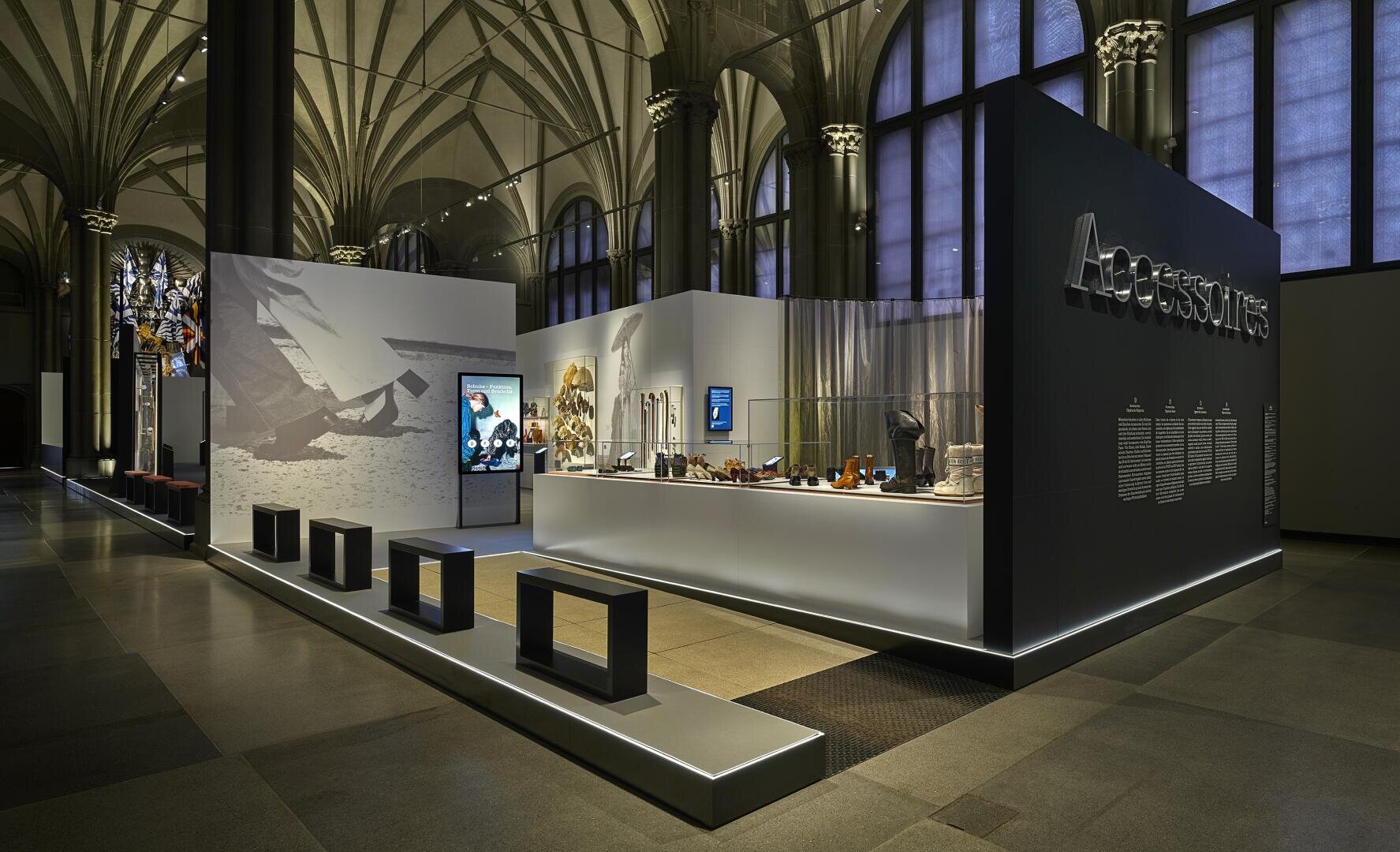 Eine moderne Museumsausstellung zeigt Accessoires in einem eleganten, gut beleuchteten Raum mit schwarzen Bänken, Glasvitrinen mit Gegenständen, interaktiven Bildschirmen und großen Wandgrafiken unter gewölbten Decken.