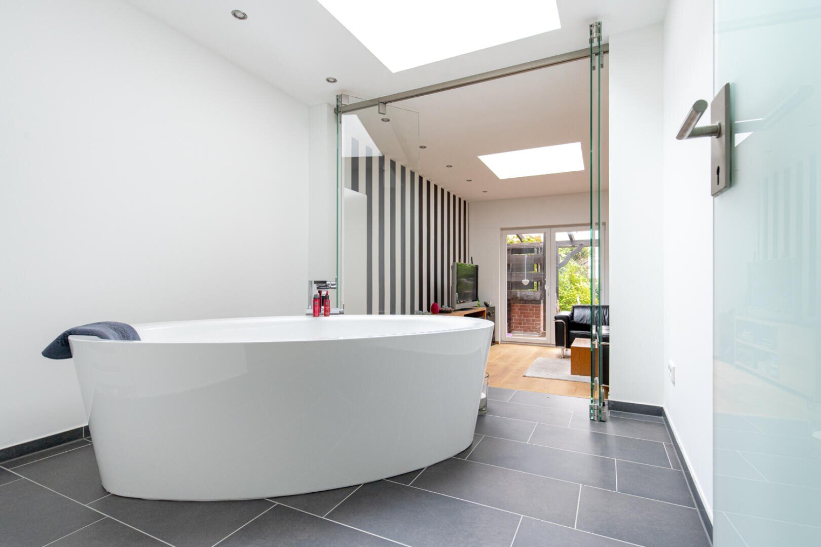 Modernes Badezimmer mit weißer freistehender Badewanne auf dunkelgrauem Fliesenboden, Glastüren zum hellen Wohnbereich mit Holzboden, TV und Terrassentüren nach draußen.