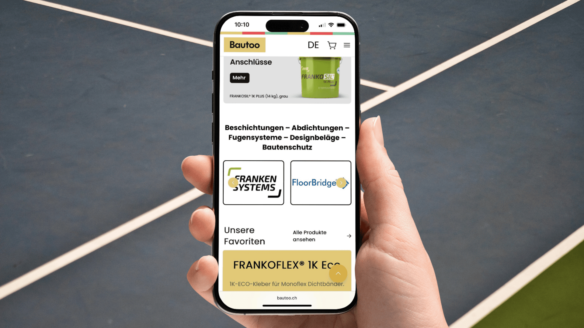 Eine Hand hält ein Smartphone, auf dem die Bautroo-Website in deutscher Sprache angezeigt wird, mit Produktkategorien und Logos auf dem Bildschirm. Im Hintergrund sind unscharfe Linien eines Tennisplatzes zu sehen.