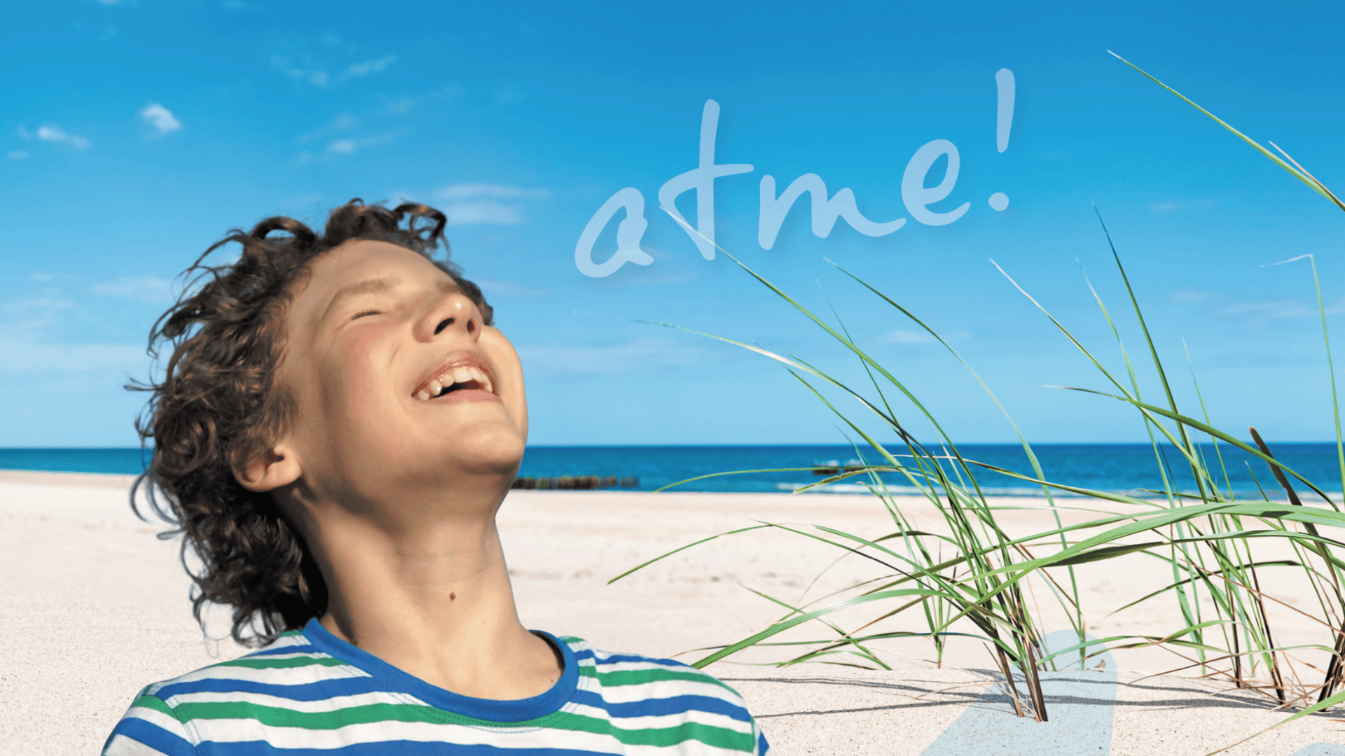 Ein fröhlicher Junge mit lockigem Haar lächelt an einem Sandstrand mit Gras und blauem Meer im Hintergrund in den Himmel. Das Wort atme! ist in den Himmel geschrieben.