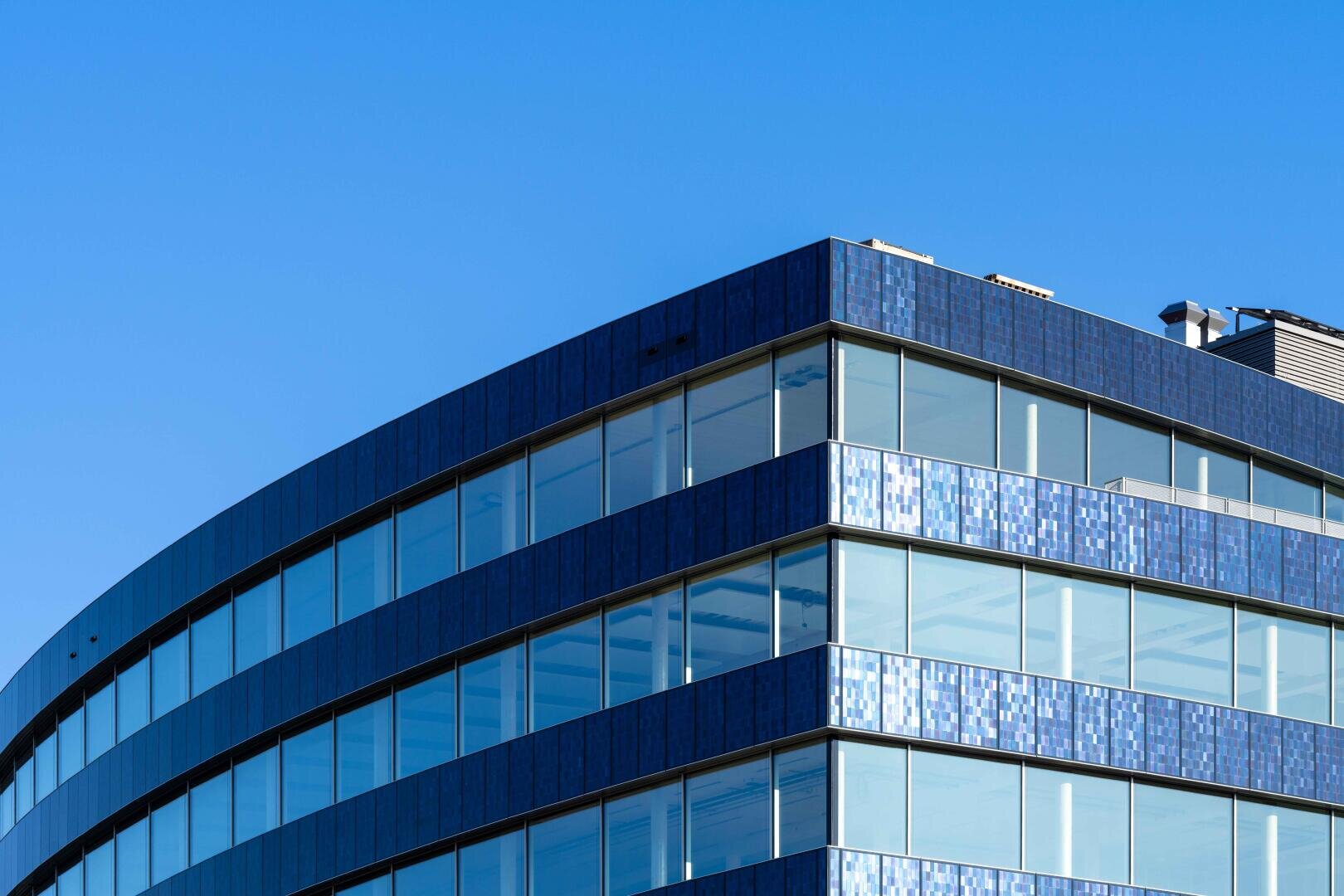 Ein modernes gläsernes Bürogebäude mit blau gekachelten Außenwänden, geschwungenen Linien und großen Fenstern vor einem klaren blauen Himmel.