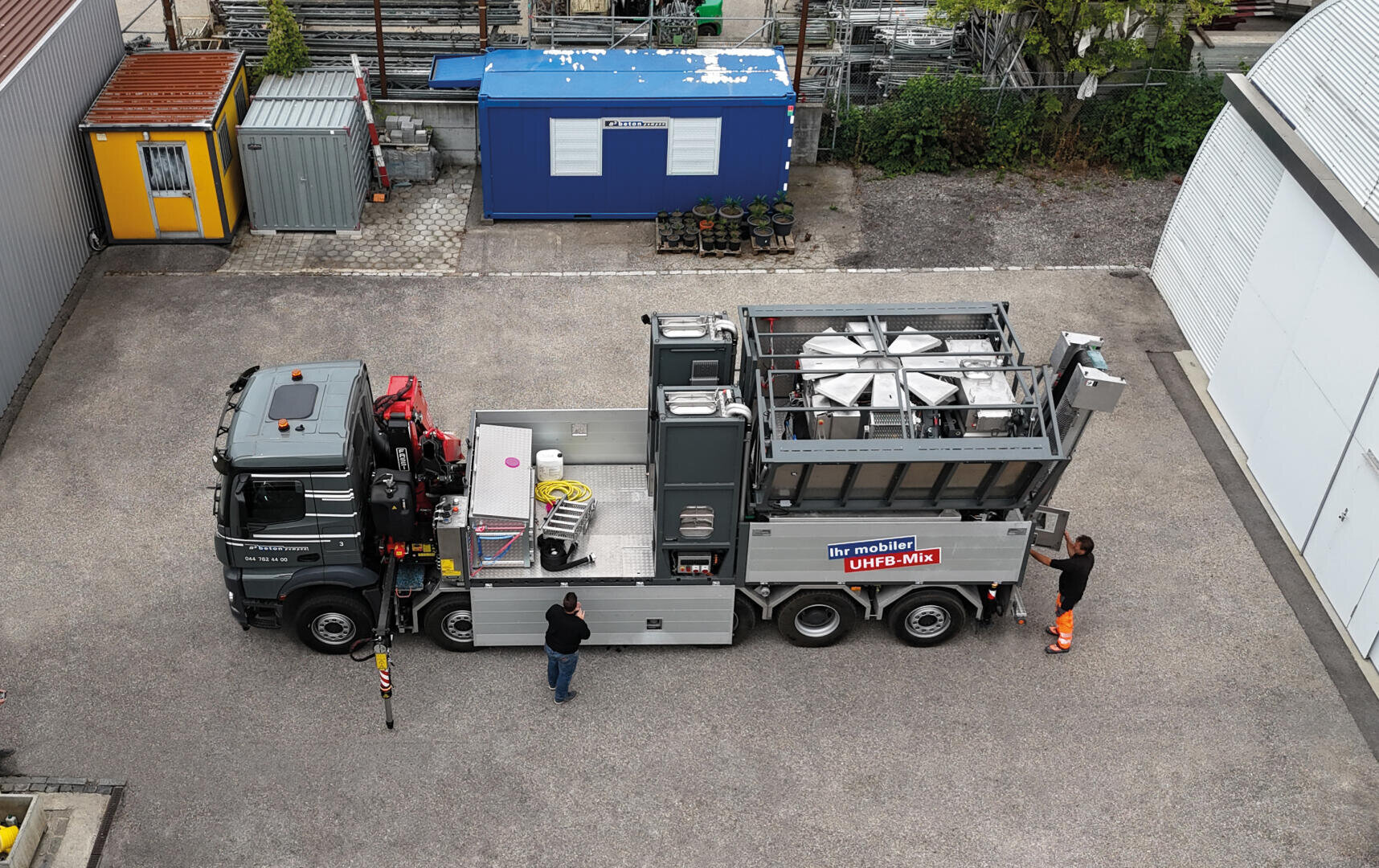 Un grand camion avec des compartiments ouverts, des machines et des équipements est garé sur un parking. Deux ouvriers se tiennent à proximité et un bâtiment portable bleu est visible à l'arrière-plan.