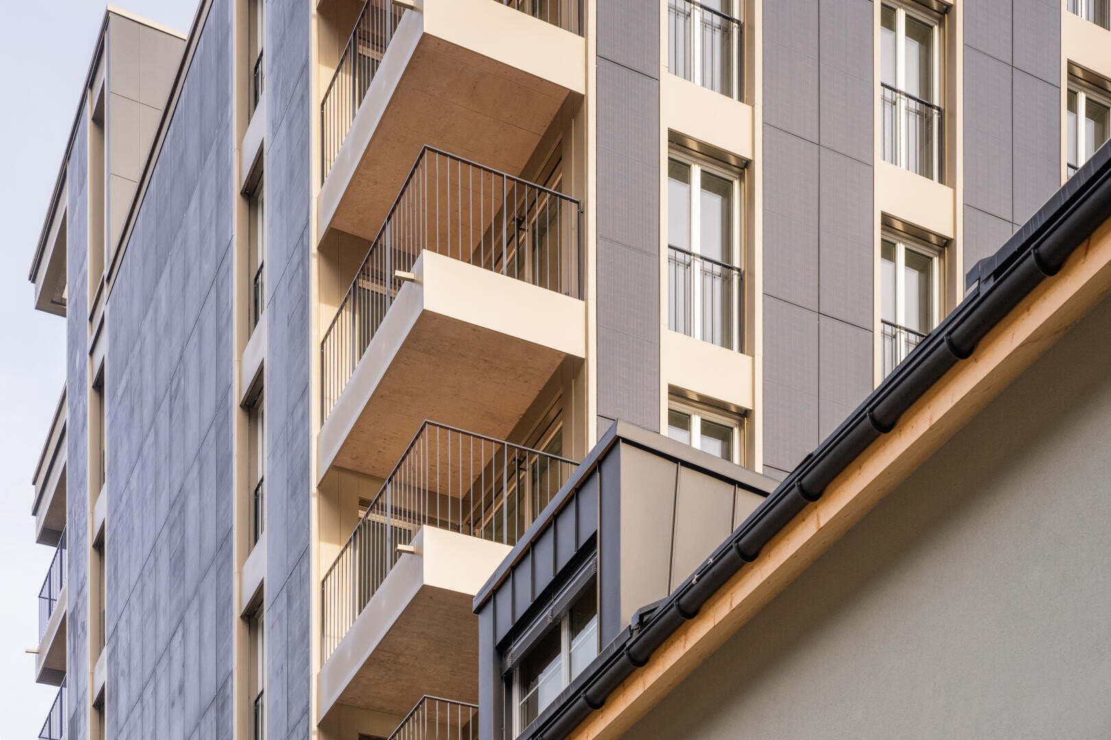 Modernes Mehrfamilienhaus mit mehreren Balkonen und großen Fenstern, von einem Winkel aus gesehen. Im Vordergrund ist ein Teil des Daches eines anderen Gebäudes zu sehen. Die Architektur zeichnet sich durch klare Linien und neutrale Farben aus.