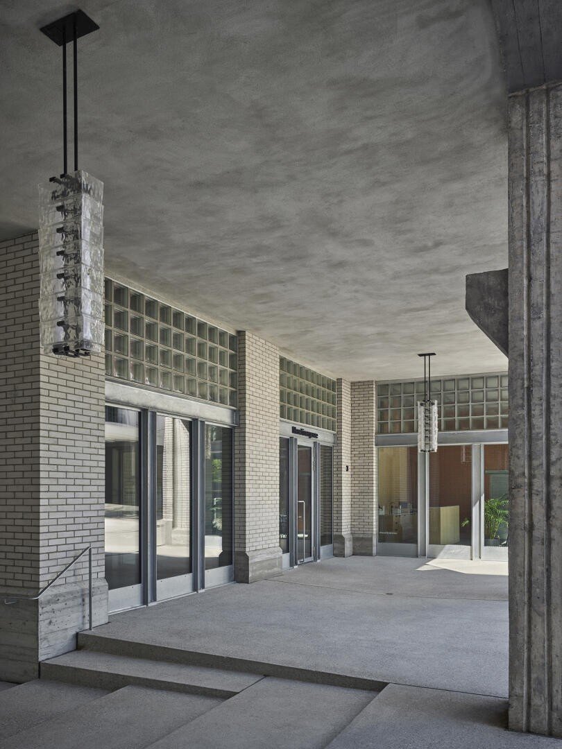 Überdachter Eingang mit Glasbausteinwänden, großen Glastüren und Betonstufen. Moderne Pendelleuchten hängen von der Decke. Der Raum hat einen minimalistischen, architektonischen Stil aus der Mitte des Jahrhunderts mit strukturierten Oberflächen und klaren Linien.