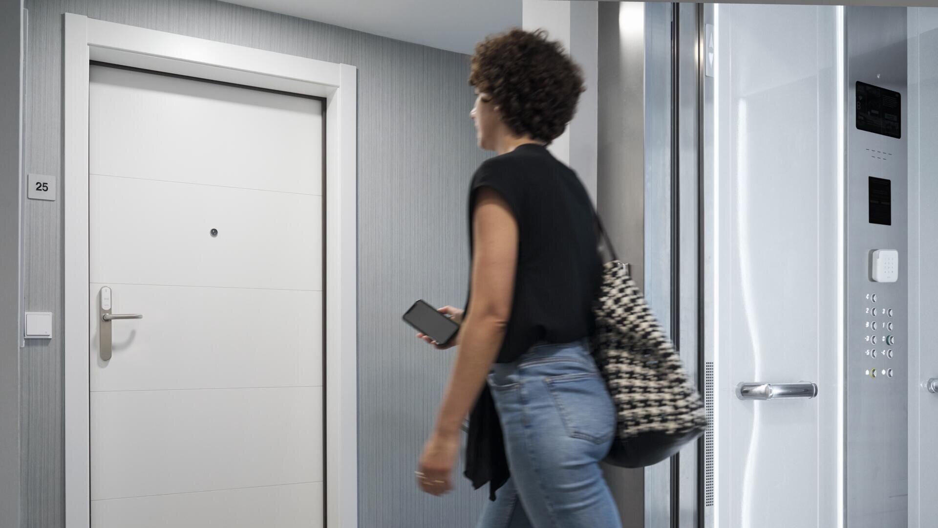 Une personne aux cheveux bouclés, portant une chemise noire et un jean bleu, tenant un téléphone à la main et portant un sac à motifs, passe devant une porte blanche et un ascenseur ouvert dans un couloir moderne.