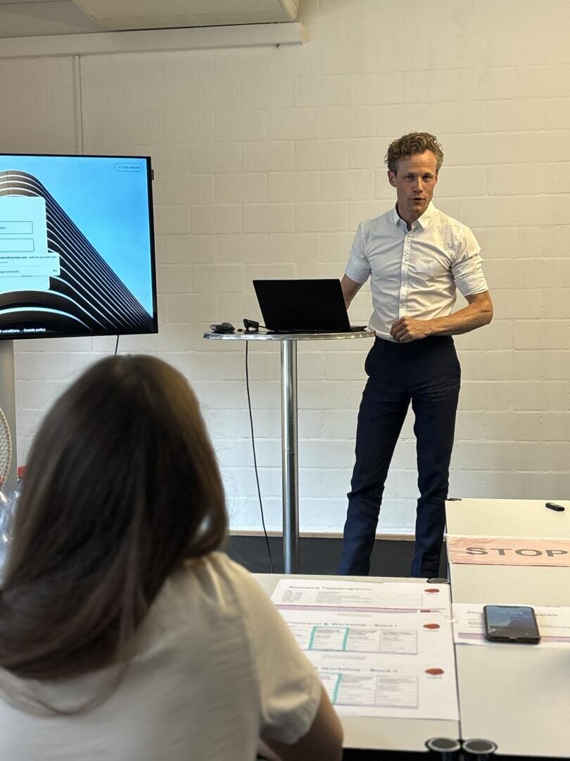 Ein Mann steht mit einem Laptop auf einem Podium und hält eine Präsentation vor einem sitzenden Publikum. Auf einem Bildschirm wird eine Webseite mit einem Architekturbild angezeigt. Auf dem Tisch im Vordergrund liegen Papiere und ein Telefon.