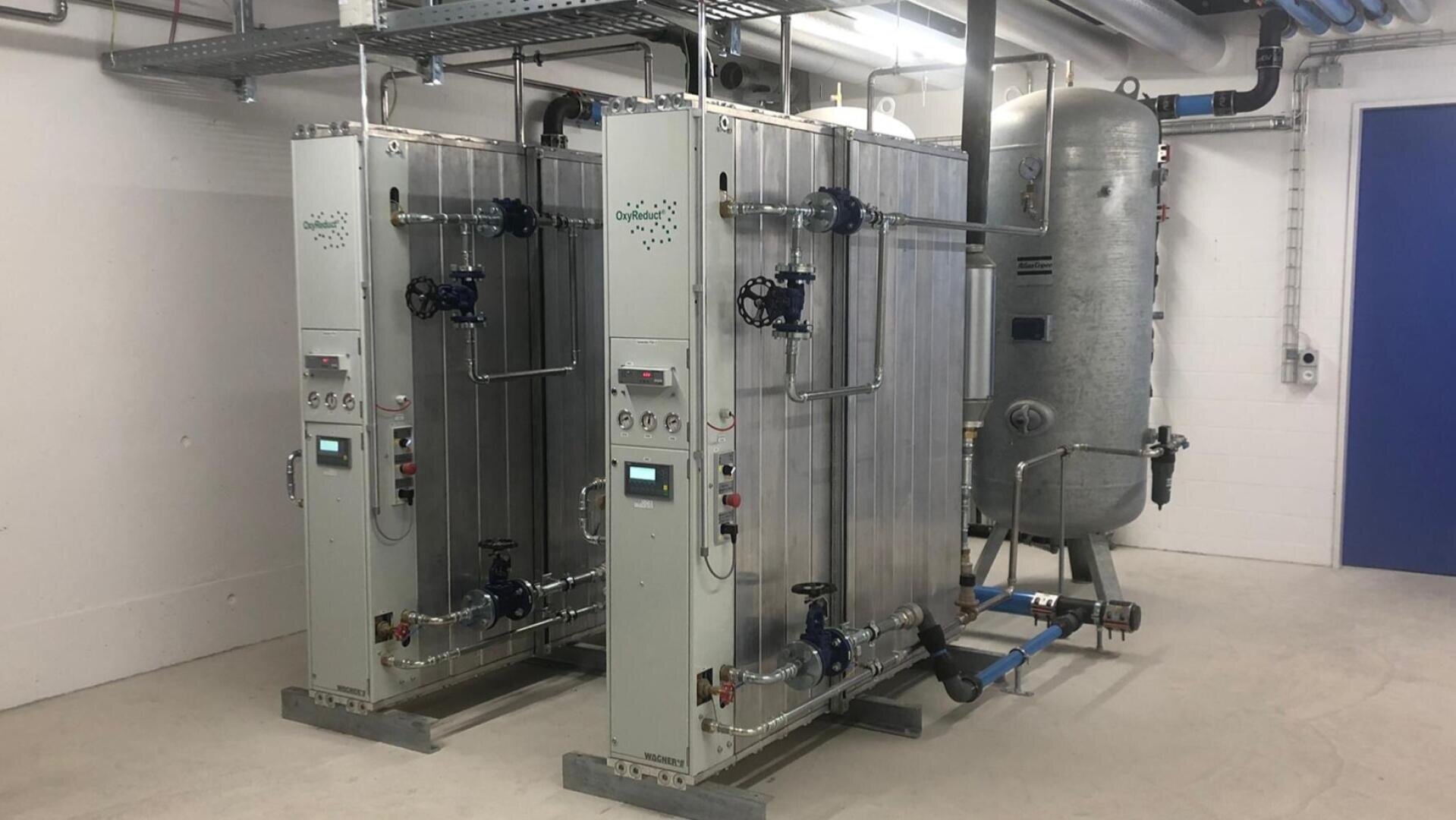 Deux grandes installations industrielles de traitement de l'eau avec des panneaux de commande et des tuyauteries sont placées à côté d'un réservoir cylindrique dans une pièce propre et blanche avec des tuyaux visibles au plafond.