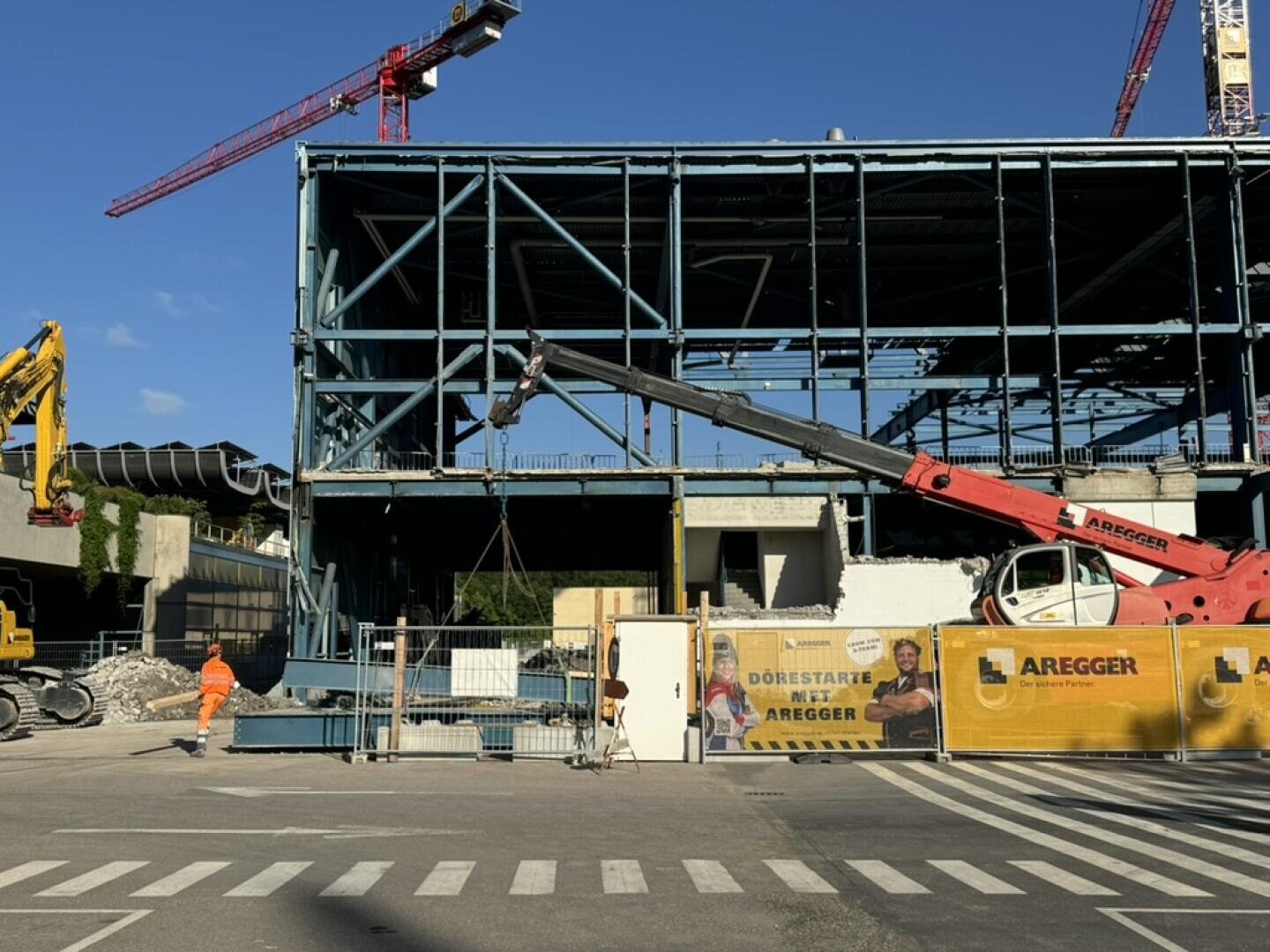 Eine Baustelle mit einem Gebäude mit Stahlrahmen, einem roten Kran und einem gelben Bagger. Ein orange gekleideter Arbeiter steht in der Nähe, und ein Banner mit dem Aregger-Logo und Arbeitern bedeckt den Zaun. Darüber ist der blaue Himmel zu sehen.