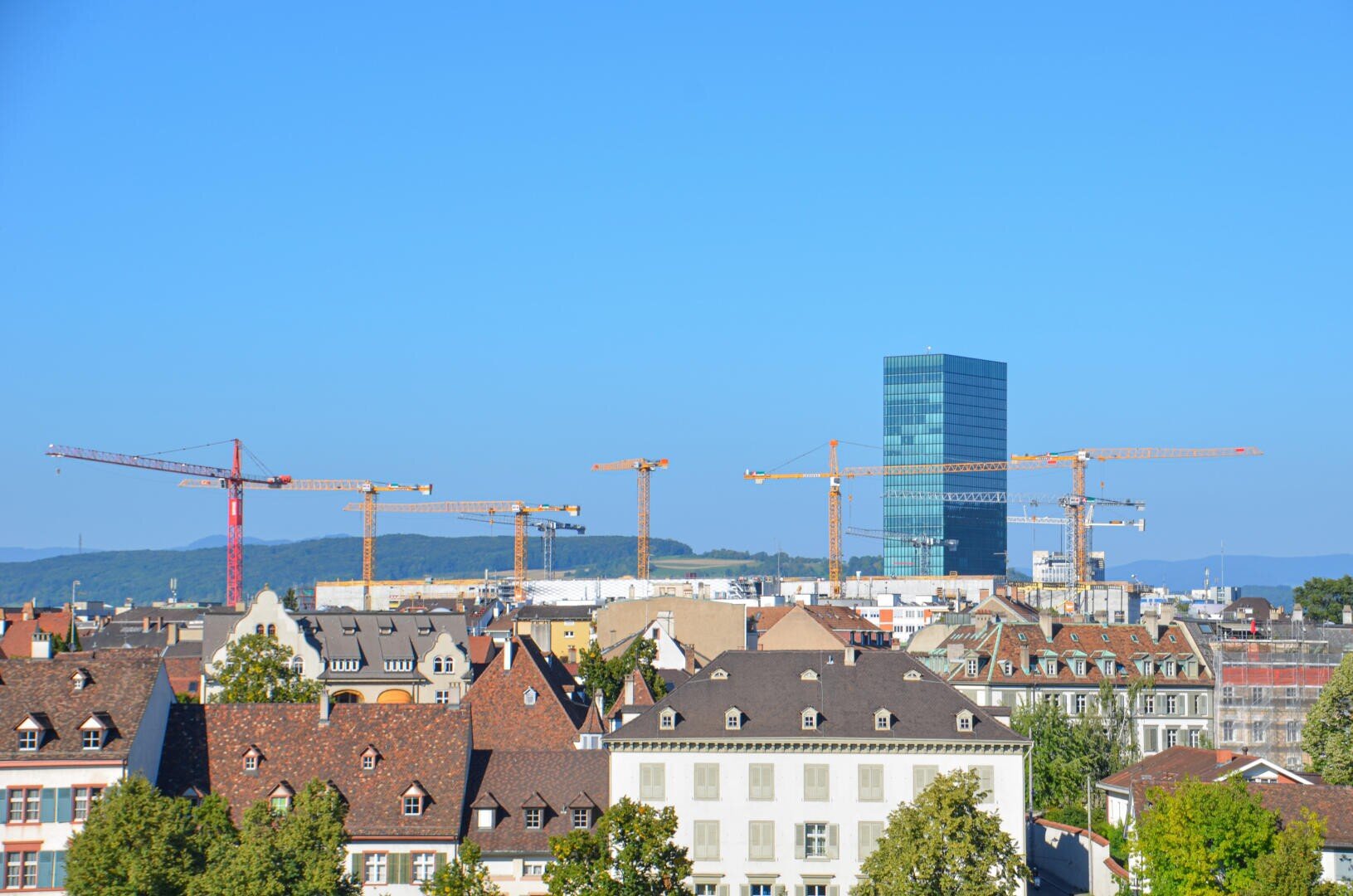Stadtbild mit traditionellen europäischen Gebäuden im Vordergrund, mehreren Baukränen in der Mitte und einem hohen modernen Glasgebäude vor einem klaren blauen Himmel im Hintergrund.