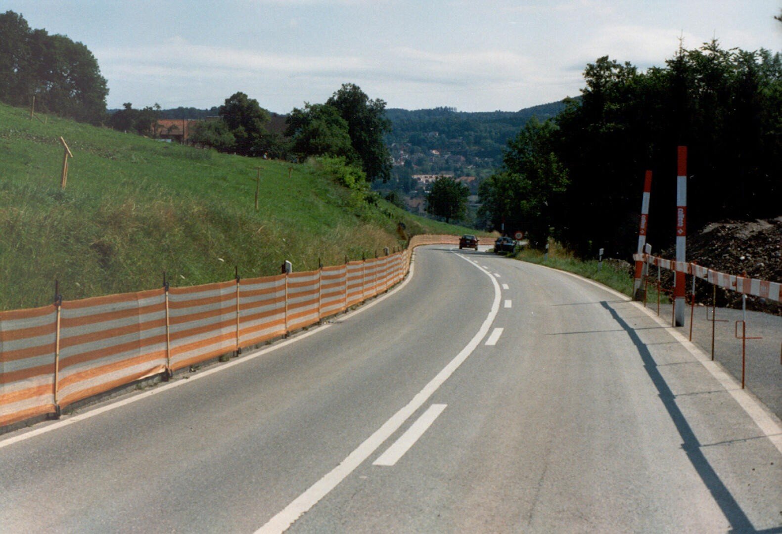 Eine kurvenreiche Landstraße mit orangefarbenen und weißen Baustellenabsperrungen auf einer Seite, gesäumt von Gras und Bäumen, mit ein paar Autos in der Ferne unter einem teilweise bewölkten Himmel.