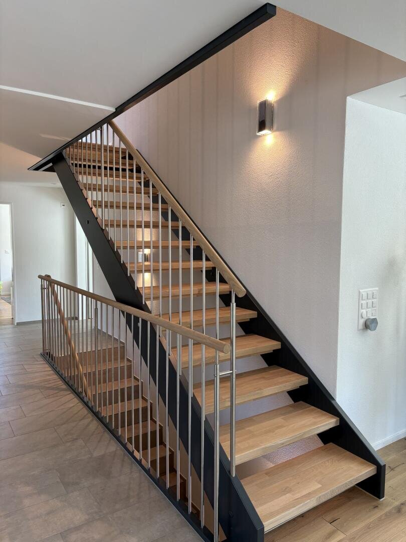 Un escalier intérieur moderne avec des marches en bois, un cadre métallique noir et une rampe verticale devant des murs blancs ; une applique murale éclaire la zone.