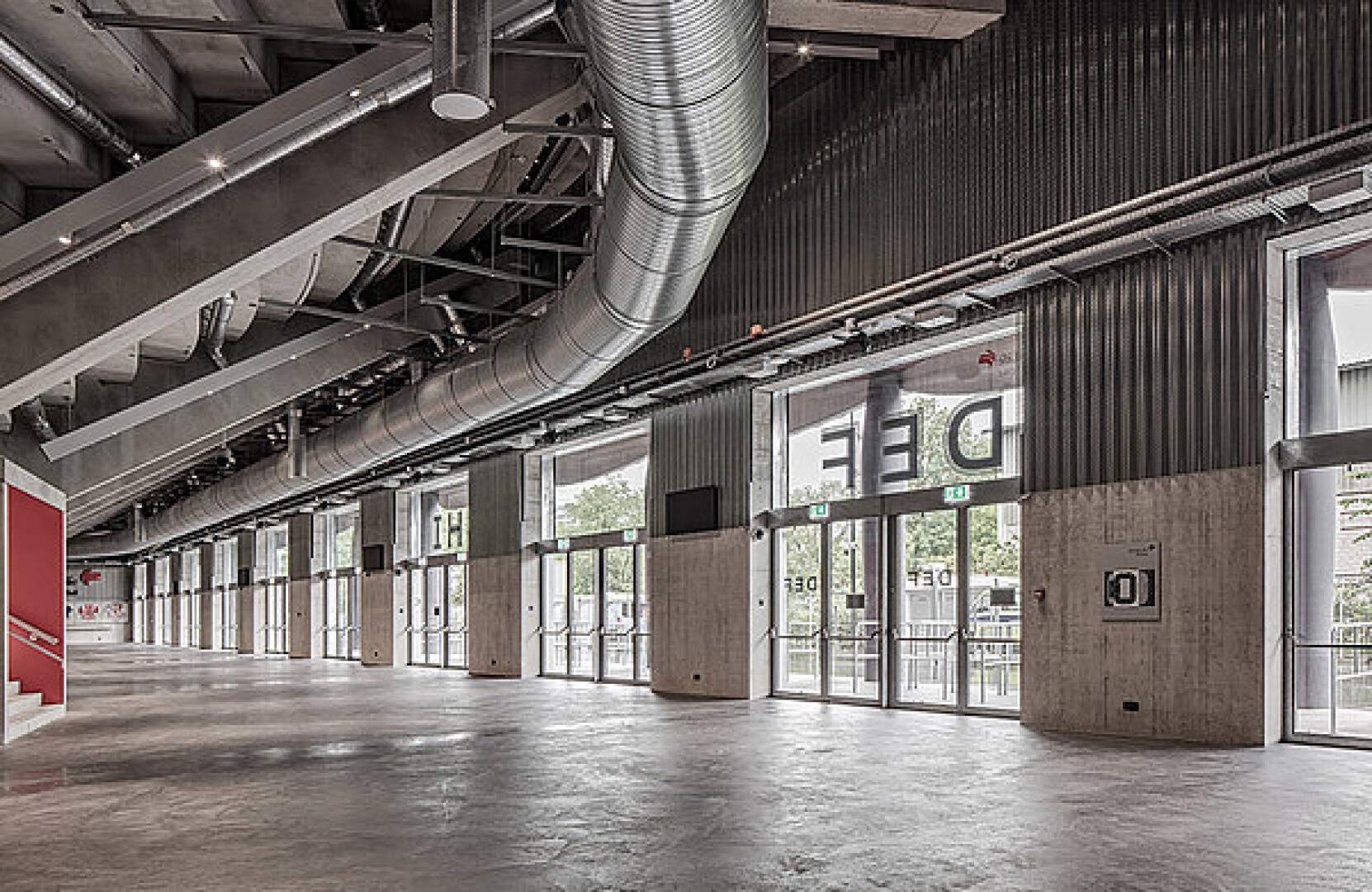 Ein geräumiger, moderner Flur mit Betonböden, großen Glastüren, freiliegenden Metallrohren an der Decke und Wänden im Industriestil. Natürliches Licht fällt durch das Glas ein, und links sind Treppen sichtbar.