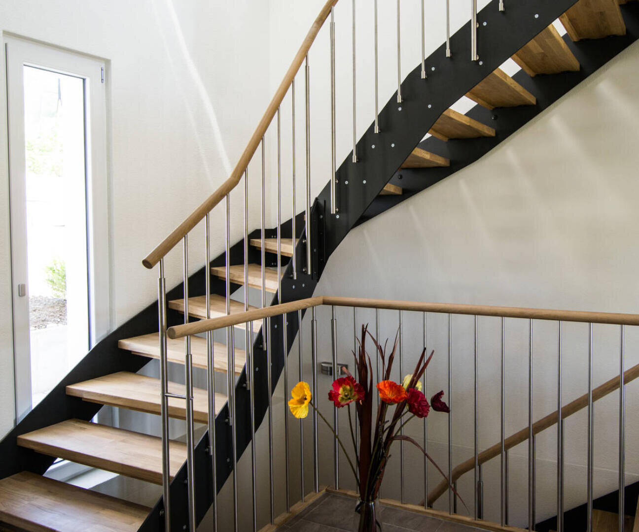 Escalier intérieur moderne avec des marches en bois, des limons métalliques noirs et des rampes métalliques. Un vase avec des fleurs artificielles colorées est posé sur une table au bas de l'escalier. La lumière naturelle entre par une porte sur la gauche.